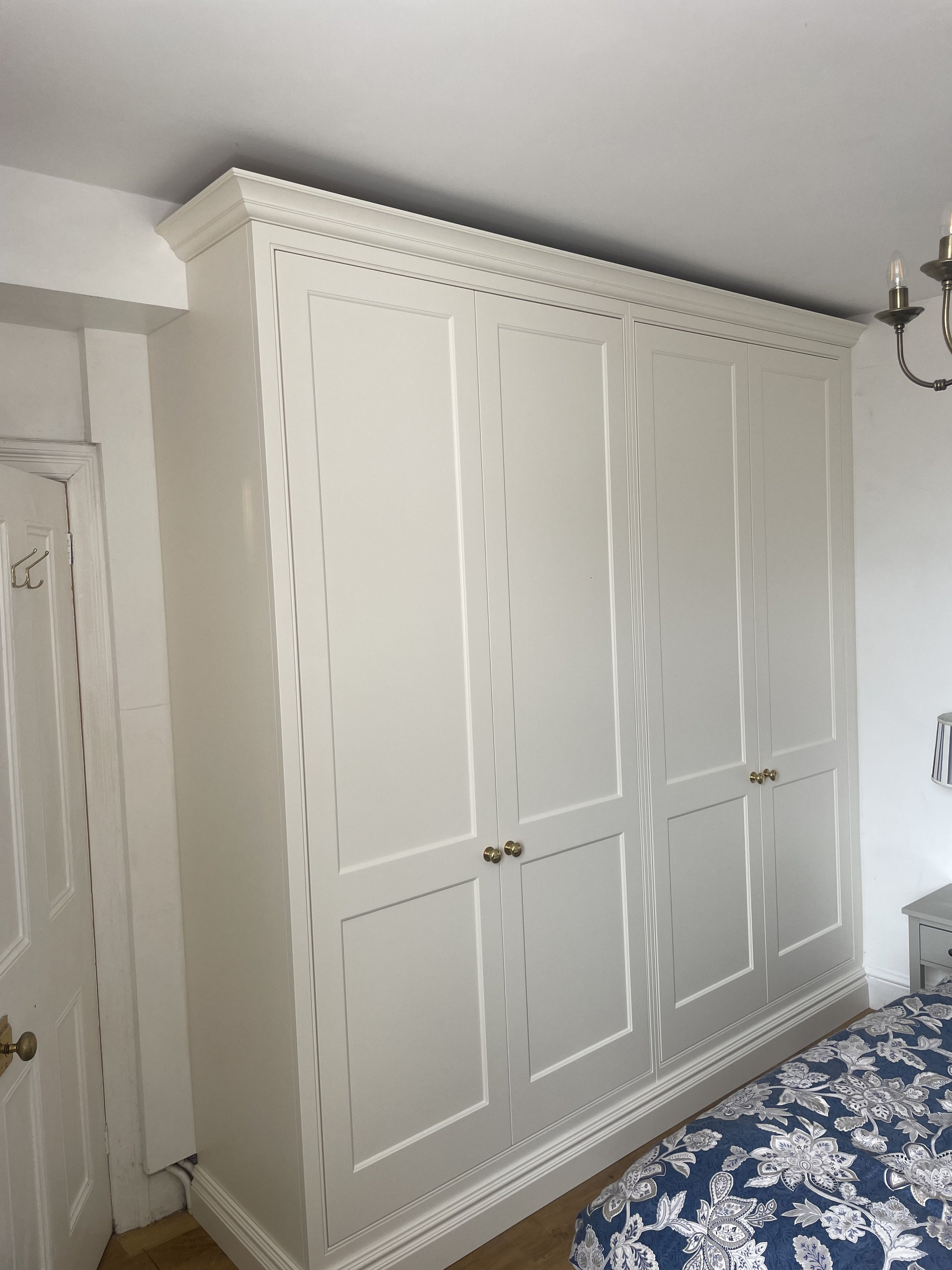 A white wardrobe with four doors and gold knobs in a bedroom. Part of a bed with a blue and white floral mattress and a small nightstand are visible.