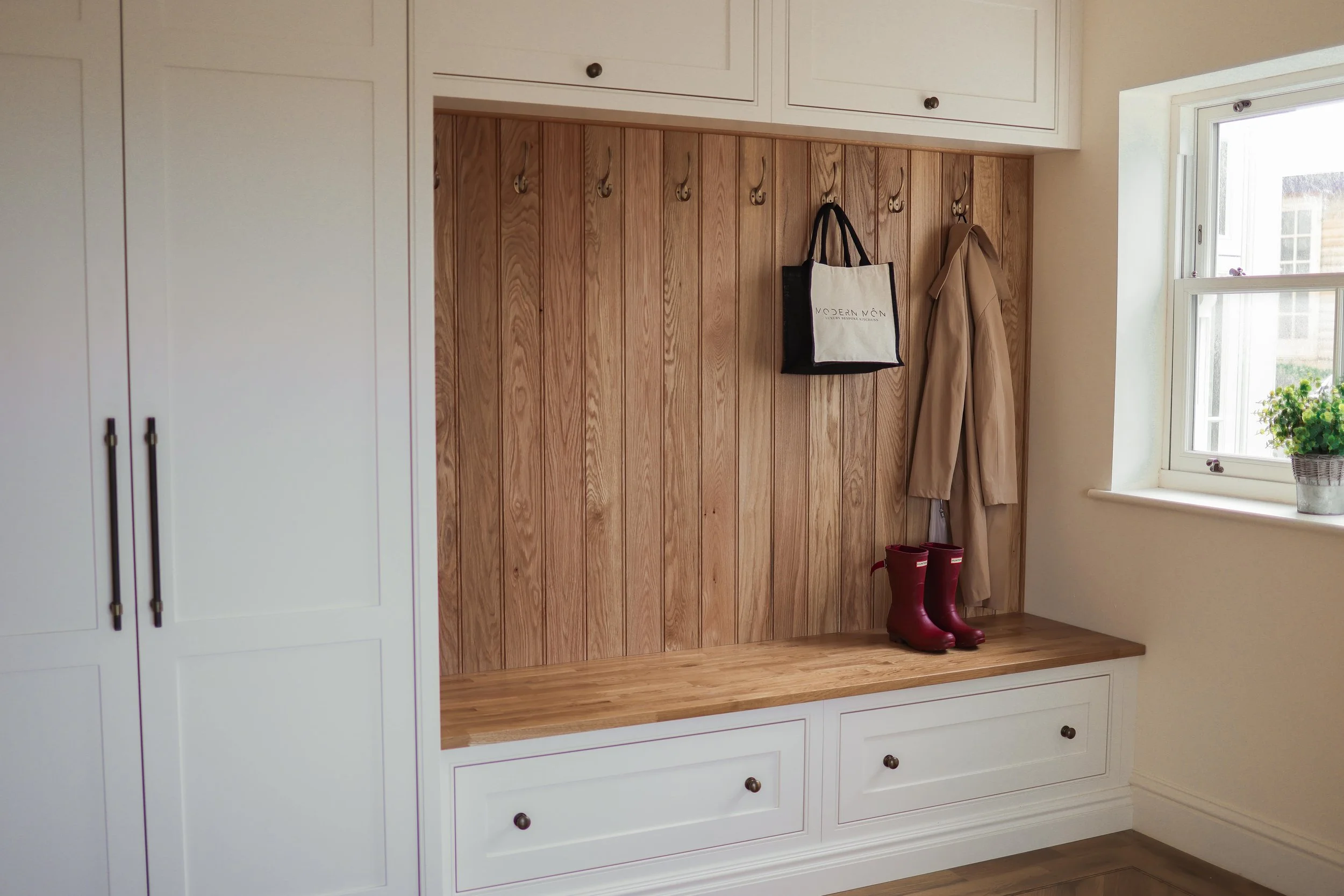 Entryway with white cabinets, wooden wall panel with hooks holding a bag and coat, red boots on bench, window with potted plant.