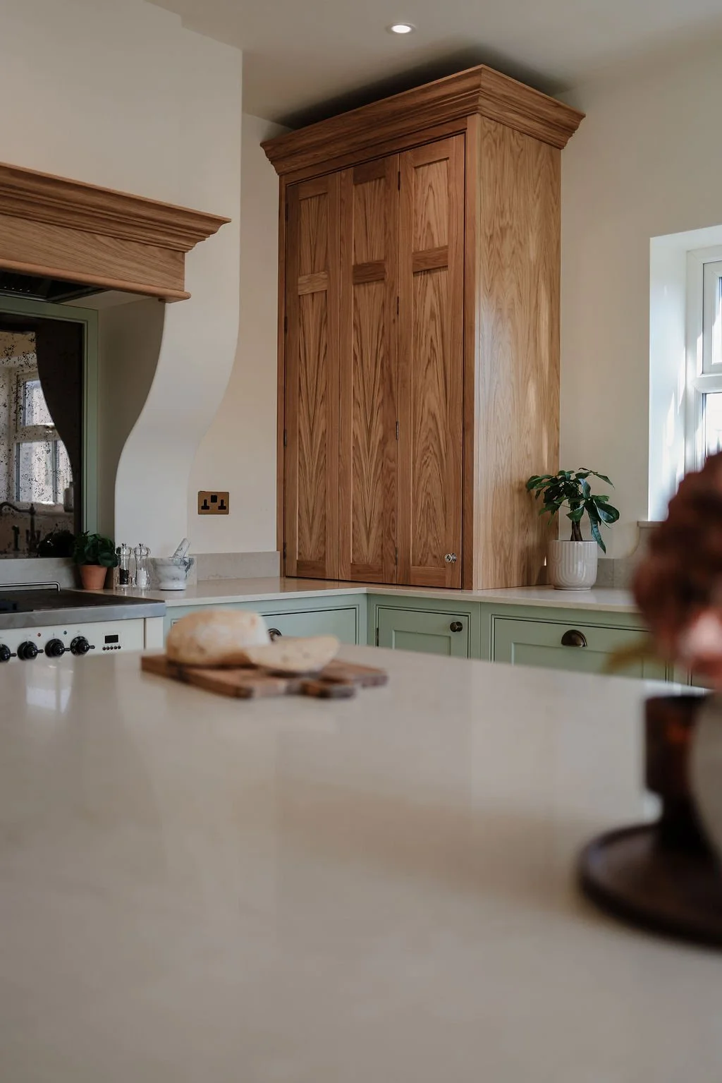 A kitchen with pastel green cabinets, a wooden corner cabinet, a window with sunlight, a potted plant, a cutting board with bread, and a blurred object in the foreground.