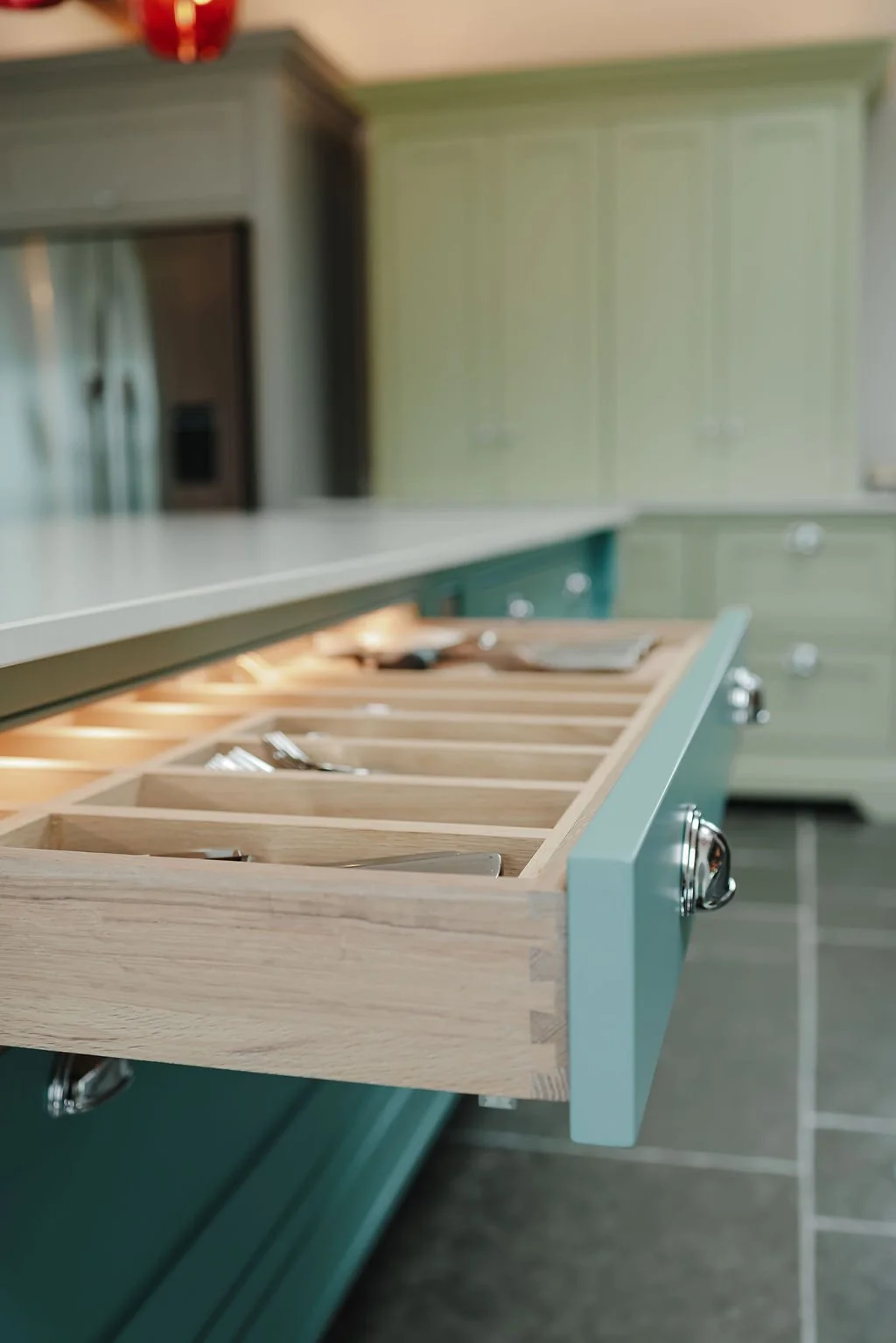 An open kitchen drawer with wooden cutlery tray containing silverware, with green cabinets and gray flooring in the background.