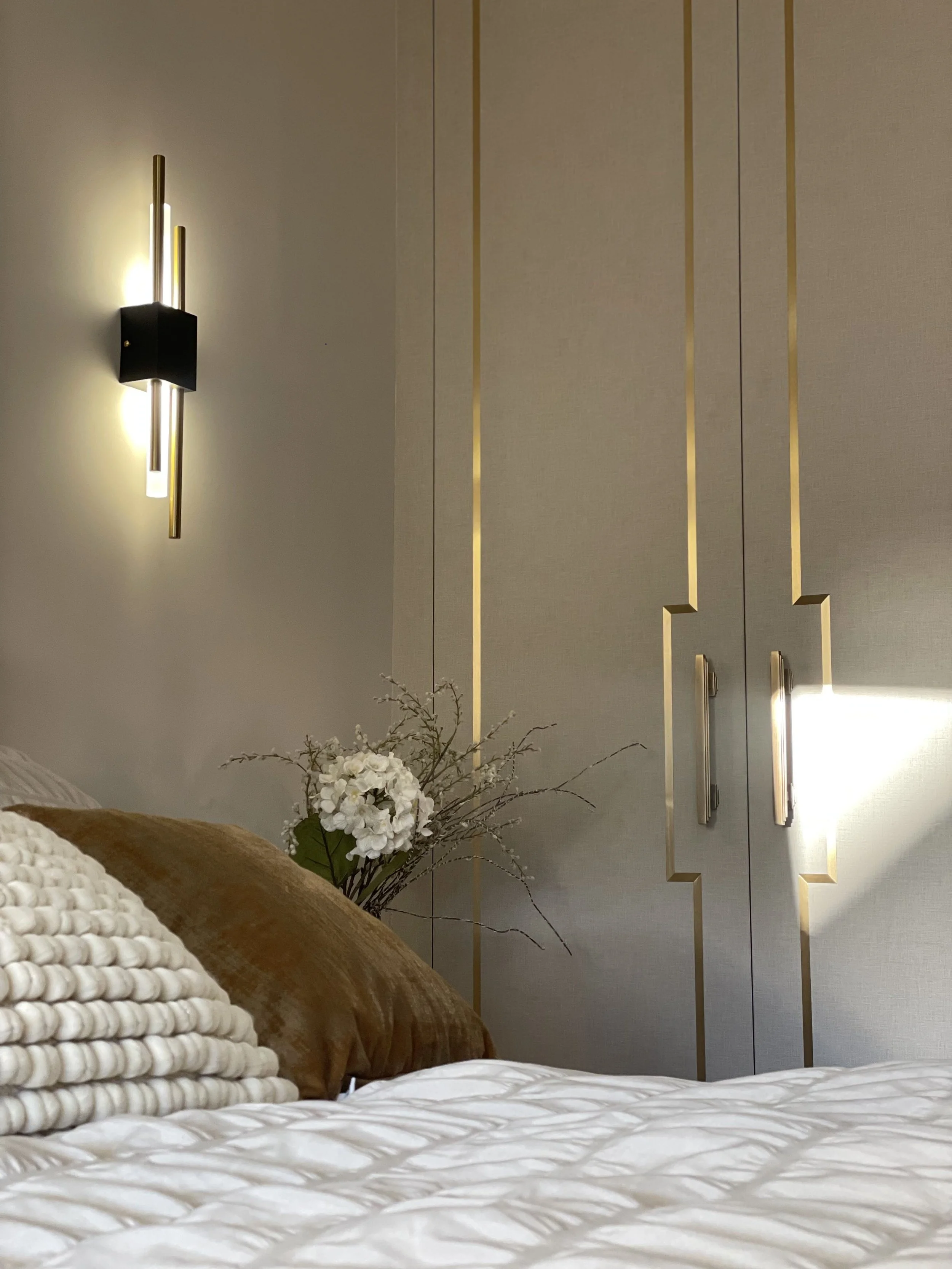 Decorative bedroom with a wall-mounted light fixture, a vase with white flowers and brown pillows, and gold accents on the wall.