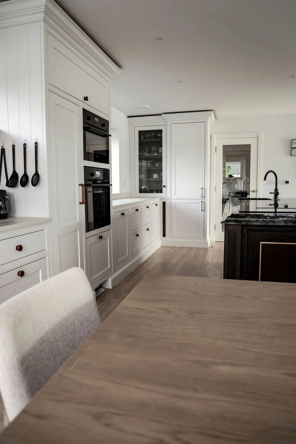 A modern kitchen with white cabinetry, black hardware, and a black marble island. The kitchen includes built-in appliances and a glass-front cabinet, with a door leading to another room and a window providing natural light.