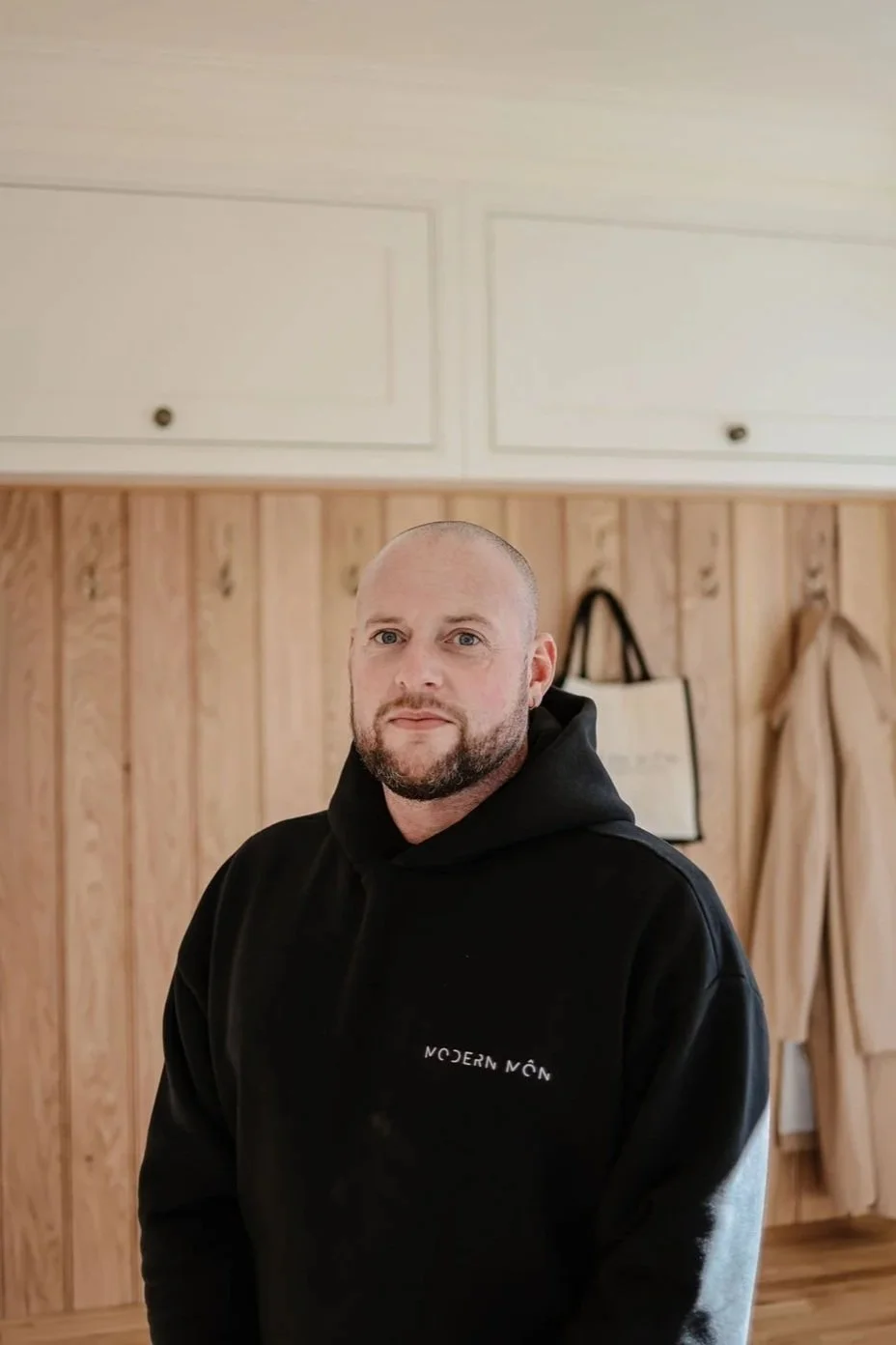 A man with a beard and shaved head wearing a black hoodie with 'MODERN NON' logo, standing in front of a wooden paneled wall with hanging items, including a beige coat and a bag.