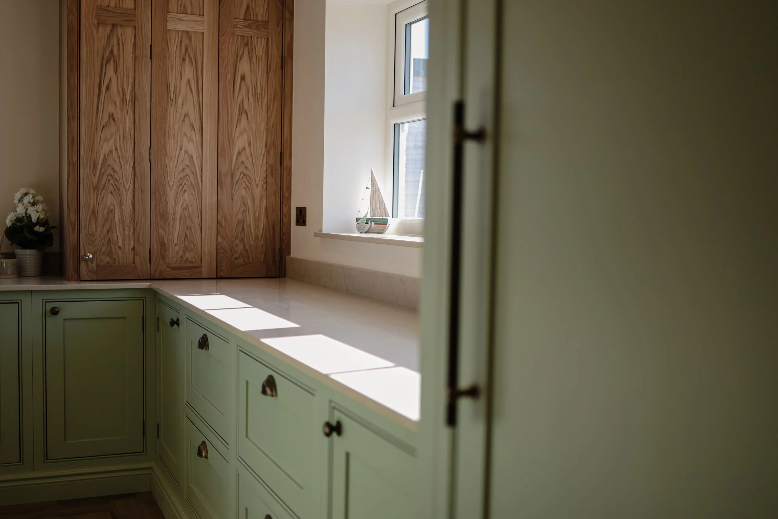 A kitchen view with pale green cabinets, a marble countertop, a window with sunlight casting shadows, and a decorative sailboat on the windowsill. There is also a brown wooden cabinet and a potted plant with white flowers.