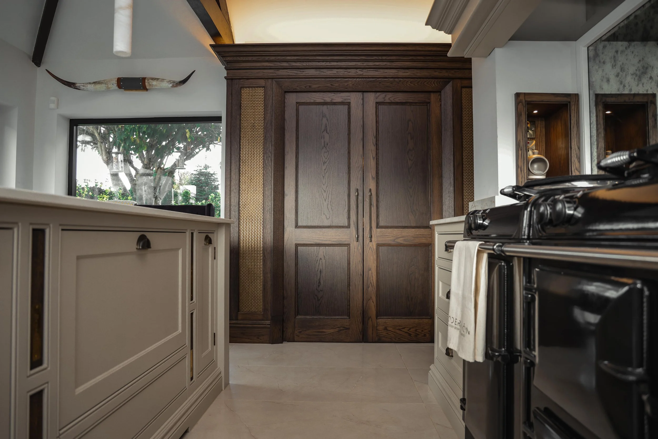 Kitchen with white cabinets, black stove, and large wooden pantry door, window showing trees outside, and a carved tusk decoration on the wall.