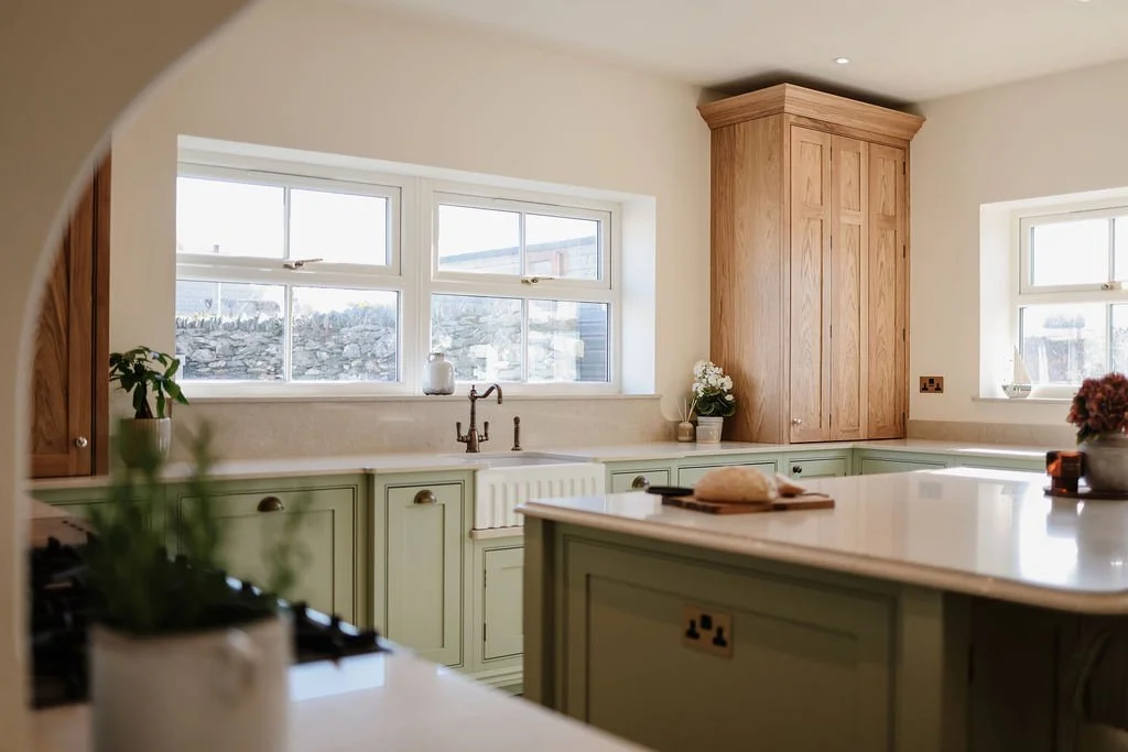 Kitchen with green cabinets, white countertops, wooden upper cabinets, two windows, plants, and decorative items.