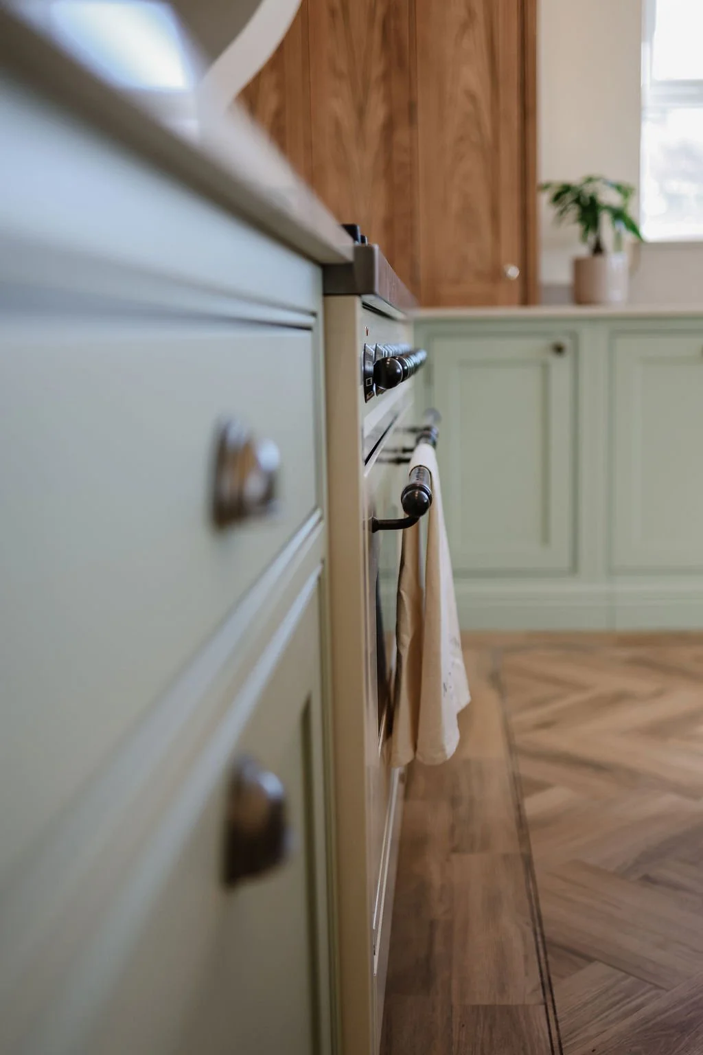 Close-up of a vintage stove with green cabinets in a kitchen, with a towel hanging from the oven handle, wooden cabinets and a window with natural light in the background.