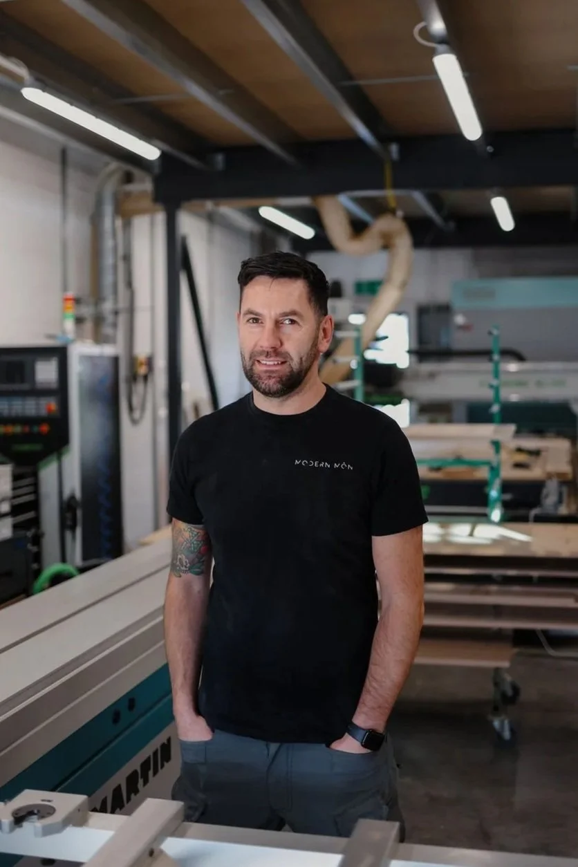 Man with dark hair and beard standing in a woodworking shop surrounded by equipment and wood projects.