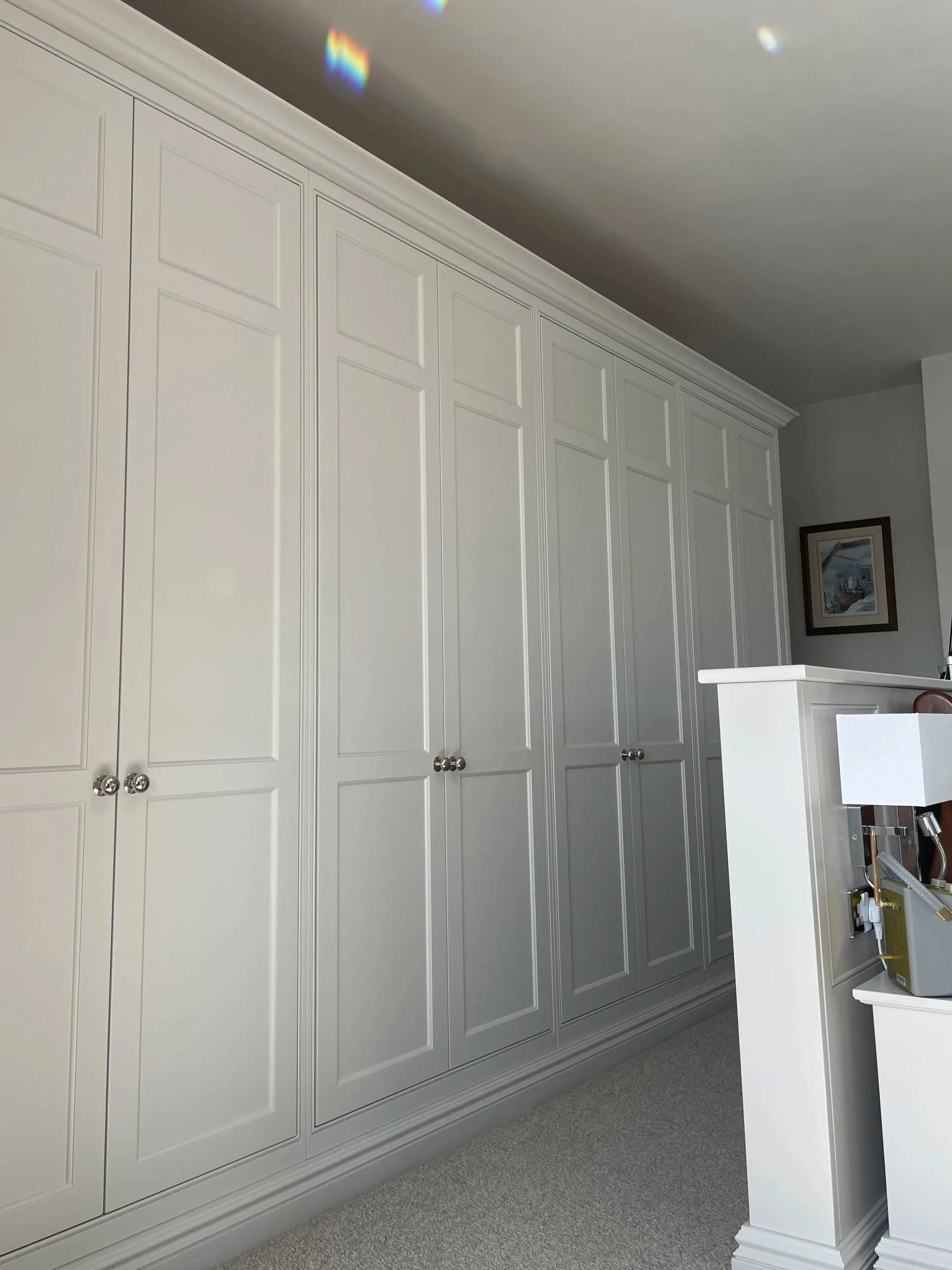 A large white closet with multiple paneled doors and shiny silver knobs in a room with gray carpet and a white curved built-in shelf or desk, with a small framed picture hanging on the wall.