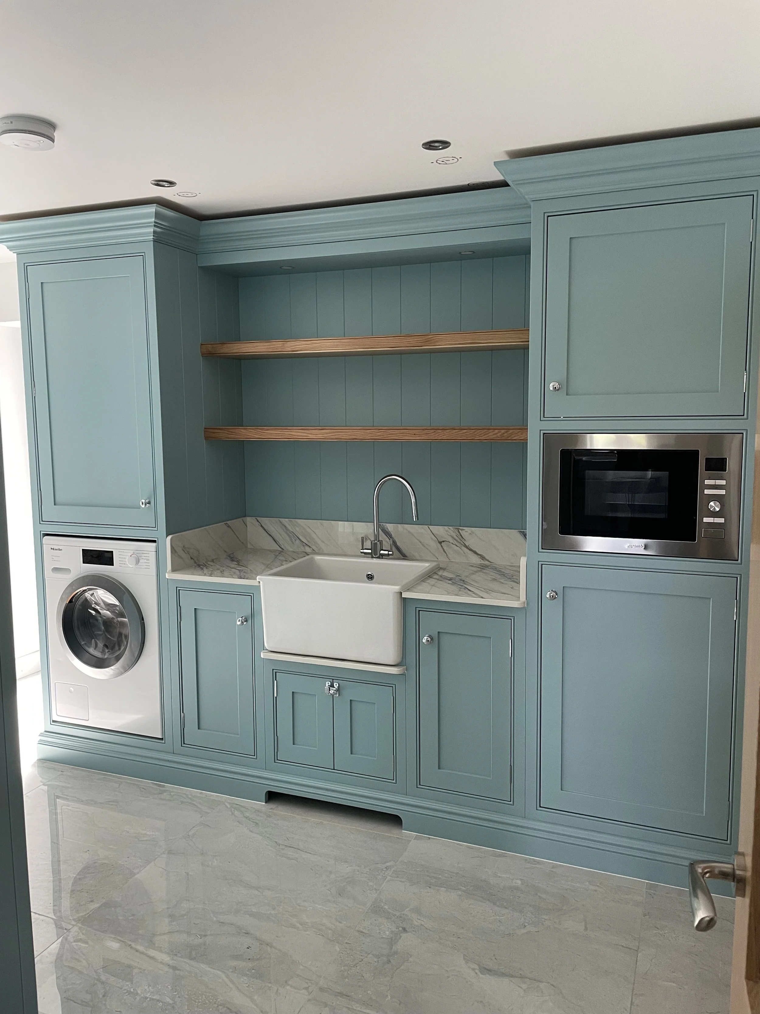 Light blue kitchen cabinets with open wooden shelves, white farmhouse sink, marble countertop, built-in microwave, washing machine, and tiled floor.