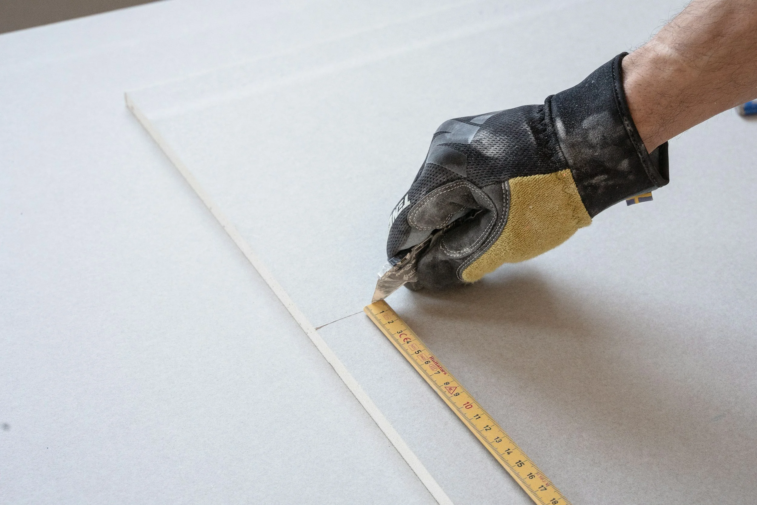 A person wearing a work glove using a measuring tape to measure a piece of white drywall.