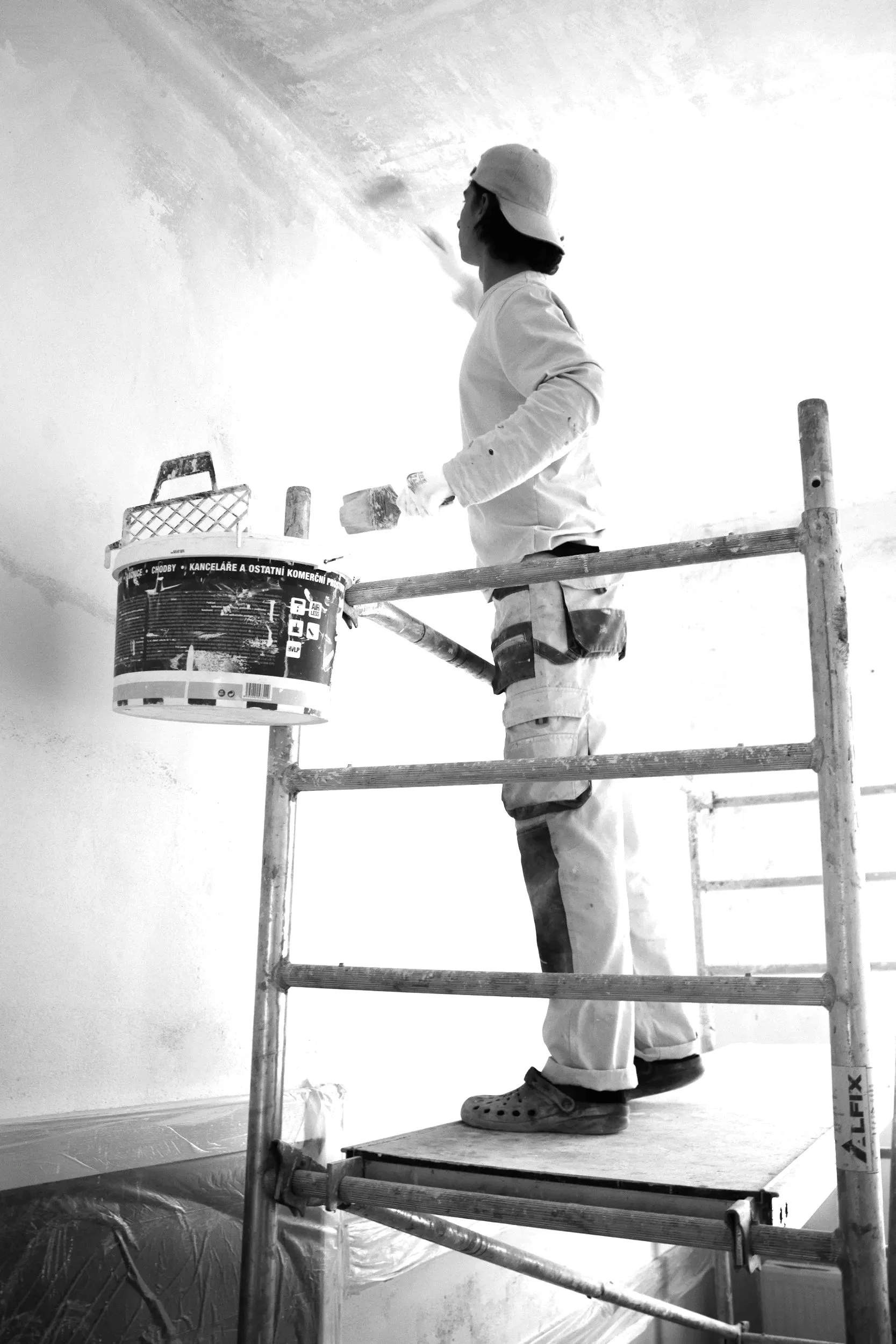 A person standing on a scaffold painting a wall with a paint roller, wearing a cap, with a paint bucket attached to the scaffolding.