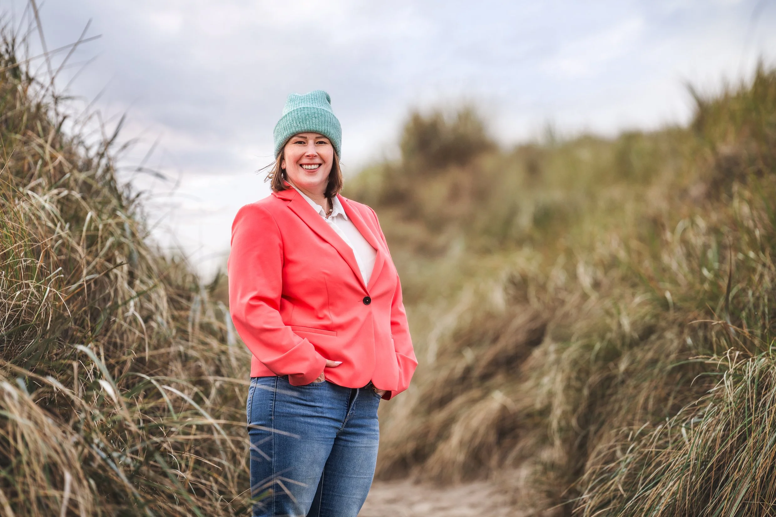 Junge Frau in rotem Blazer und grauem Hut steht zwischen Gräsern an einem sandigen Weg und lächelt in die Kamera.