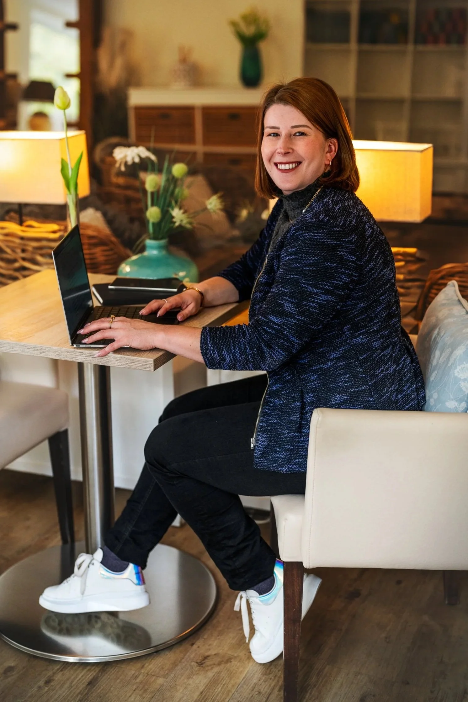 Eine lachende Frau sitzt an einem Tisch in einem gemütlichen Raum, arbeitet an einem Laptop, trägt eine dunkelblaue Jacke, schwarze Hosen und weiße Turnschuhe.