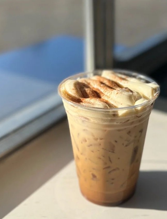 A cold coffee beverage with whipped cream and a sprinkle of cocoa powder on top, served in a clear plastic cup near a window.
