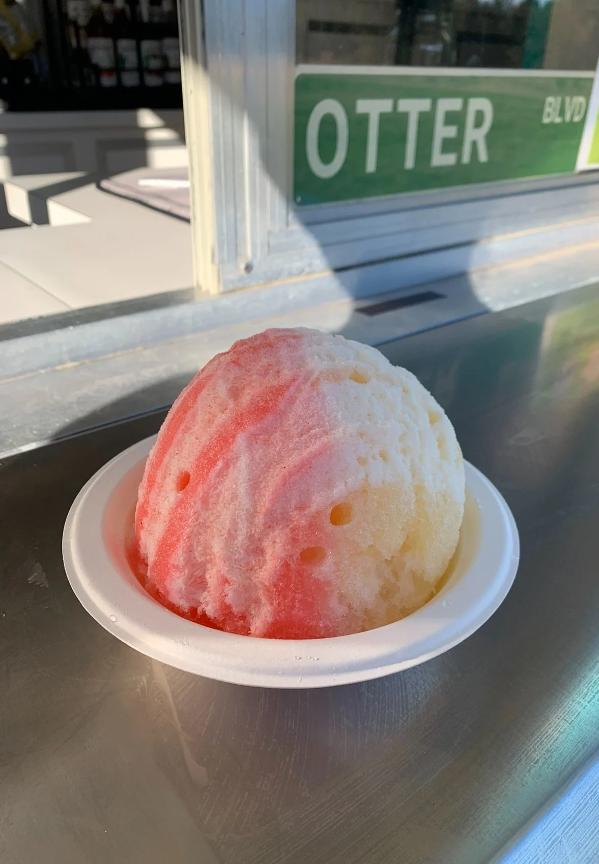 A bowl of snow cone ice with red and yellow syrup, placed on a stainless-steel surface outside a building with a green street sign that reads 'Otter Blvd'.