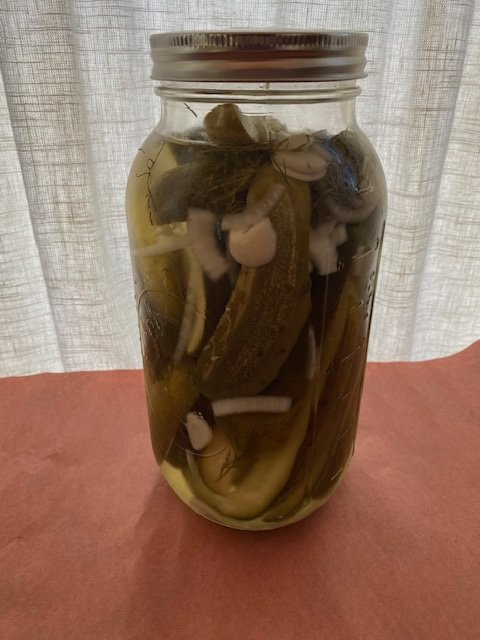 A glass jar filled with pickles submerged in liquid, with various pickle slices and garlic cloves visible inside.
