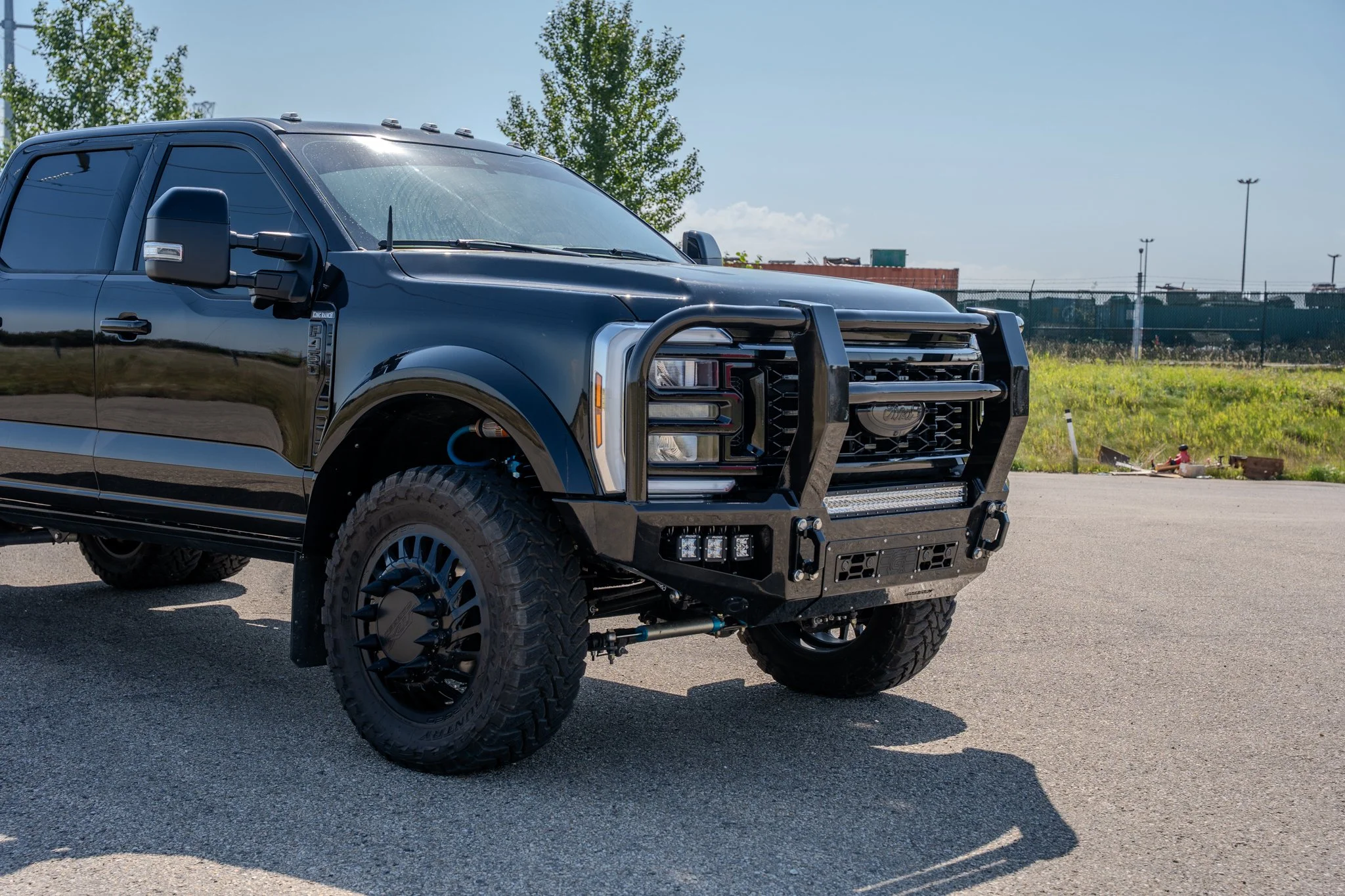 Black pickup truck with heavy-duty front bumper and grille guard parked on asphalt, green grass, and fence in the background under a clear sky. wraps. signs, branding
