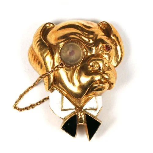Gold-tone brooch in the shape of a dog's face with an eyepatch, wearing a bow tie, and a small chain attached.