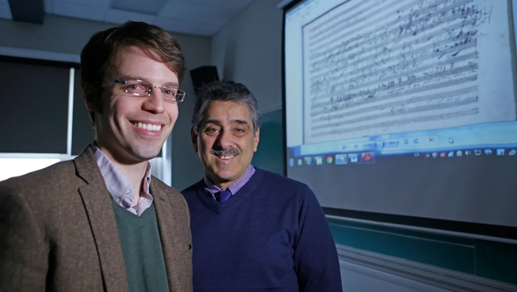 Two men smiling in front of a projector screen displaying a document or presentation in a classroom or office setting.