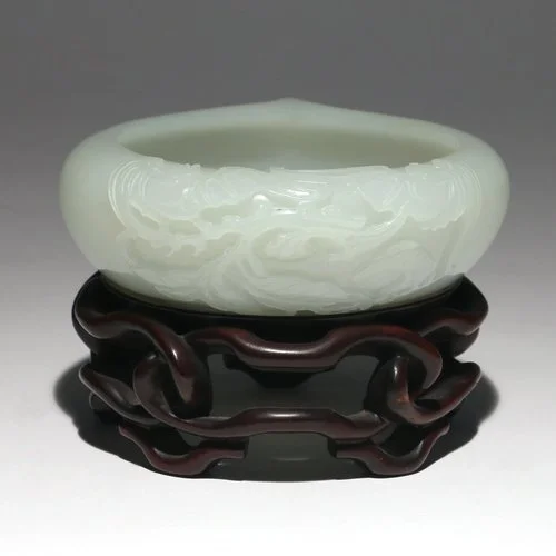CHINESE CARVED PALE JADE WATER COUPE 
low-form carved pale jade water, one side carved and decorated with two bats among leafy branches, the opposite side with a formed carved spout, resting upon a conforming openwork carved wood stand; weight of jad