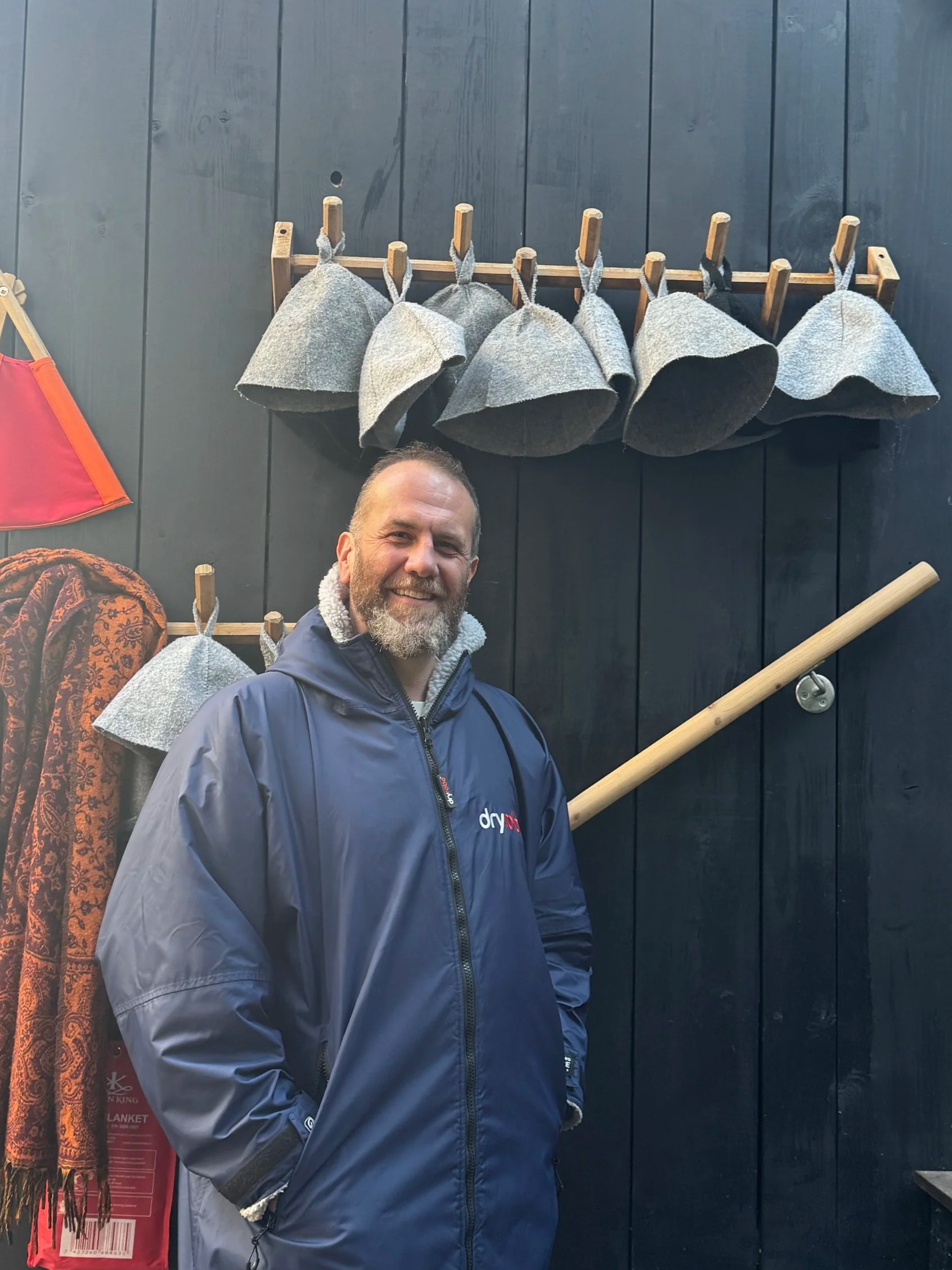Man standing outside in front of a black wall with hanging grey hats and a wooden pole with a shelf. He is wearing a dark blue waterproof jacket and smiling.