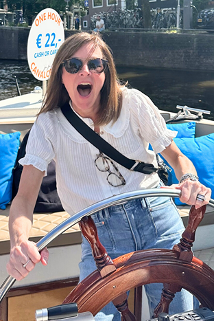 Woman with sunglasses and a white blouse steering a boat, with a sign in the background.