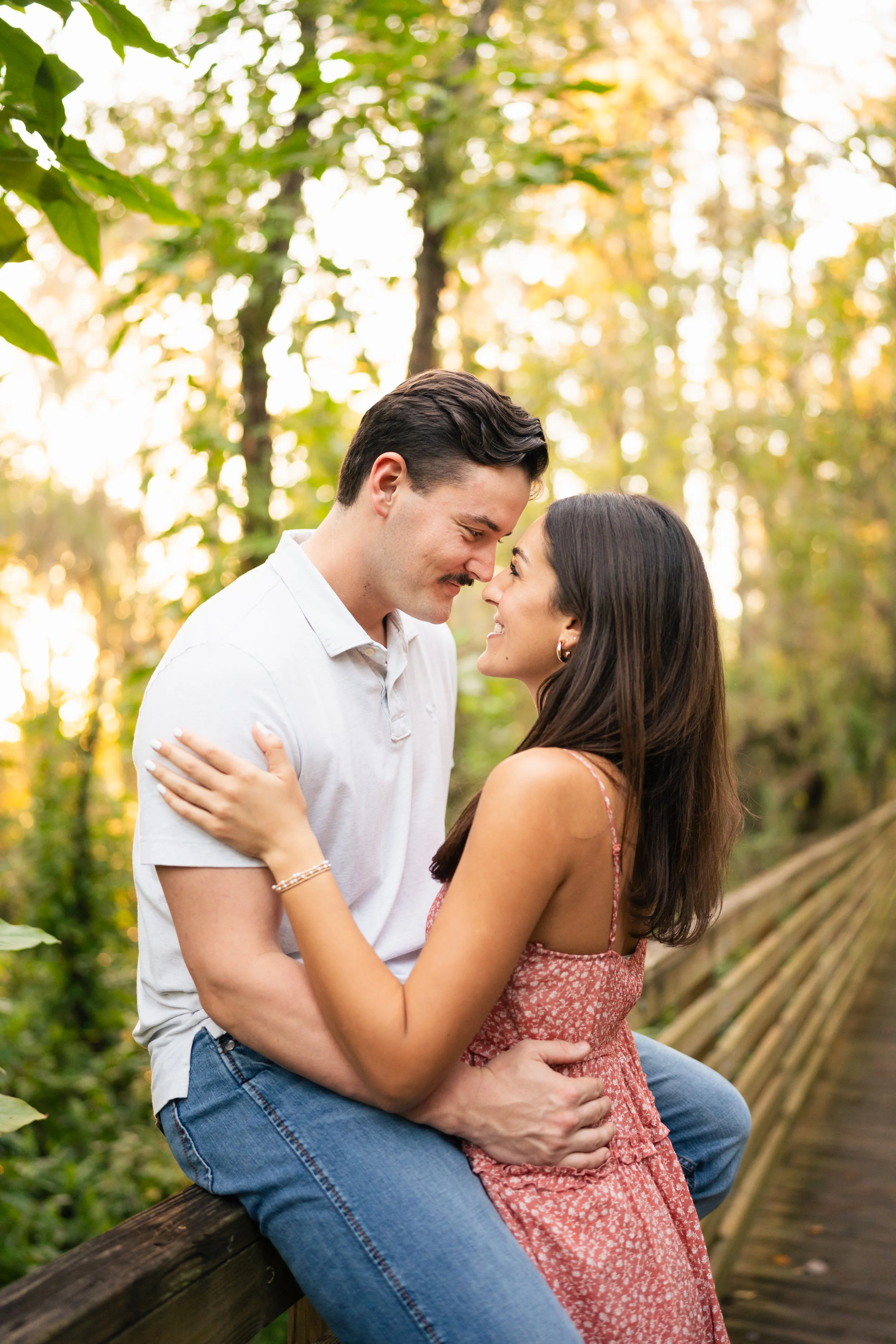 A couple sitting on a wooden railing in a forest, smiling at each other with foreheads almost touching, surrounded by green and yellow trees during sunset.
