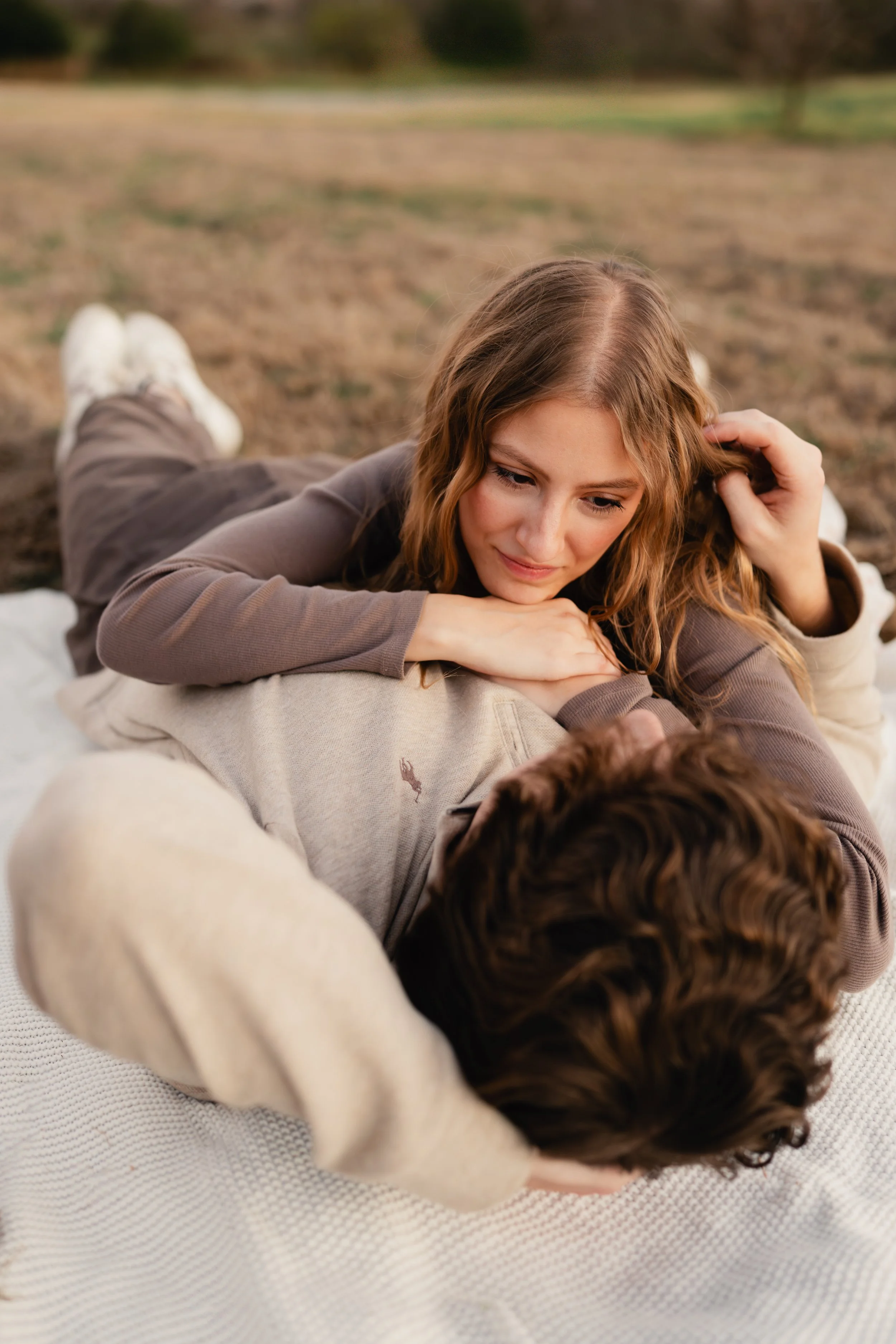 A young woman with wavy light brown hair lying on top of a person with curly dark hair outdoors on a blanket. They are on a grassy field with trees in the background. The woman is looking at the person she is on top of with a gentle expression.