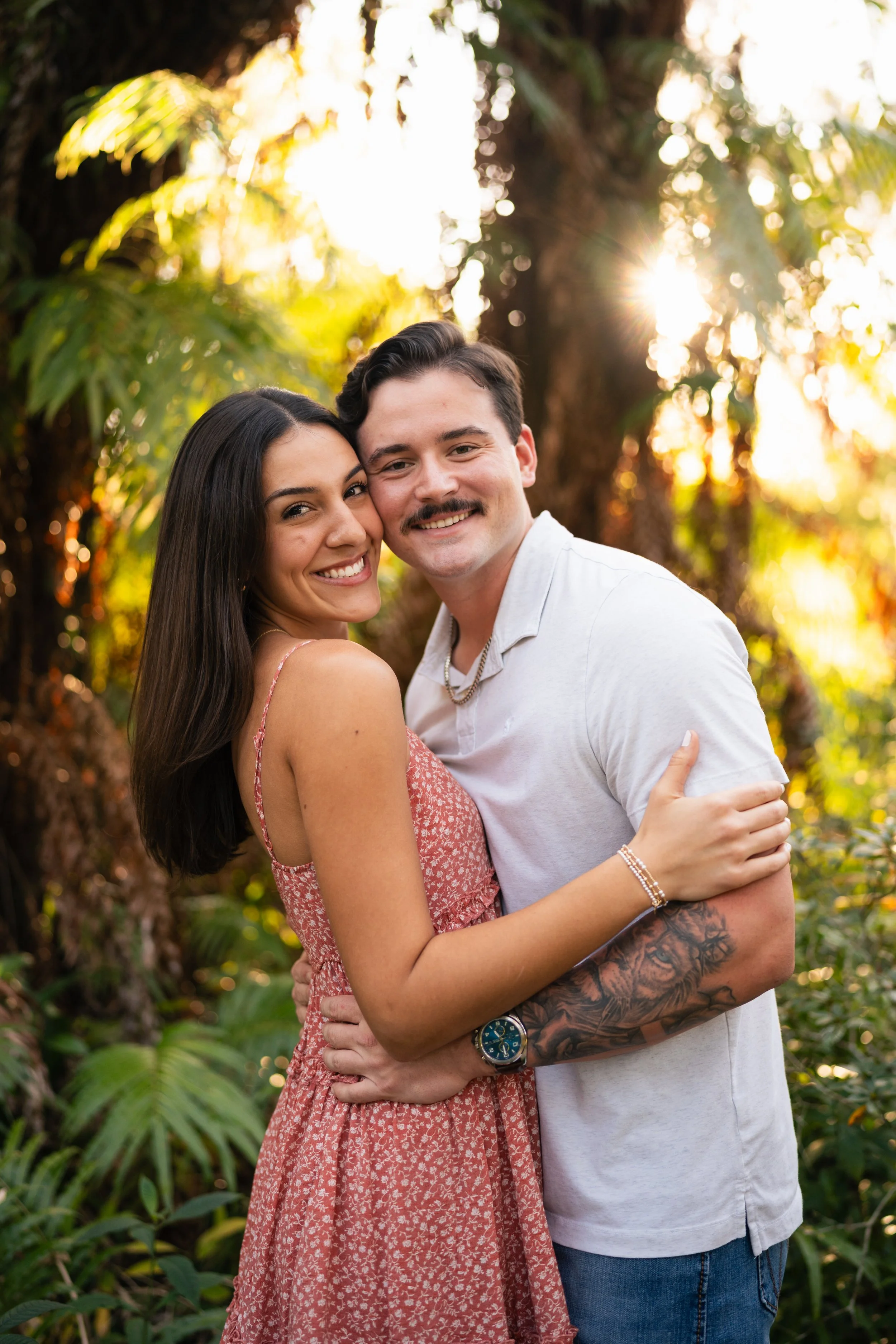 A smiling couple hugging outdoors in a sunlit forest, with green foliage and tall trees in the background.