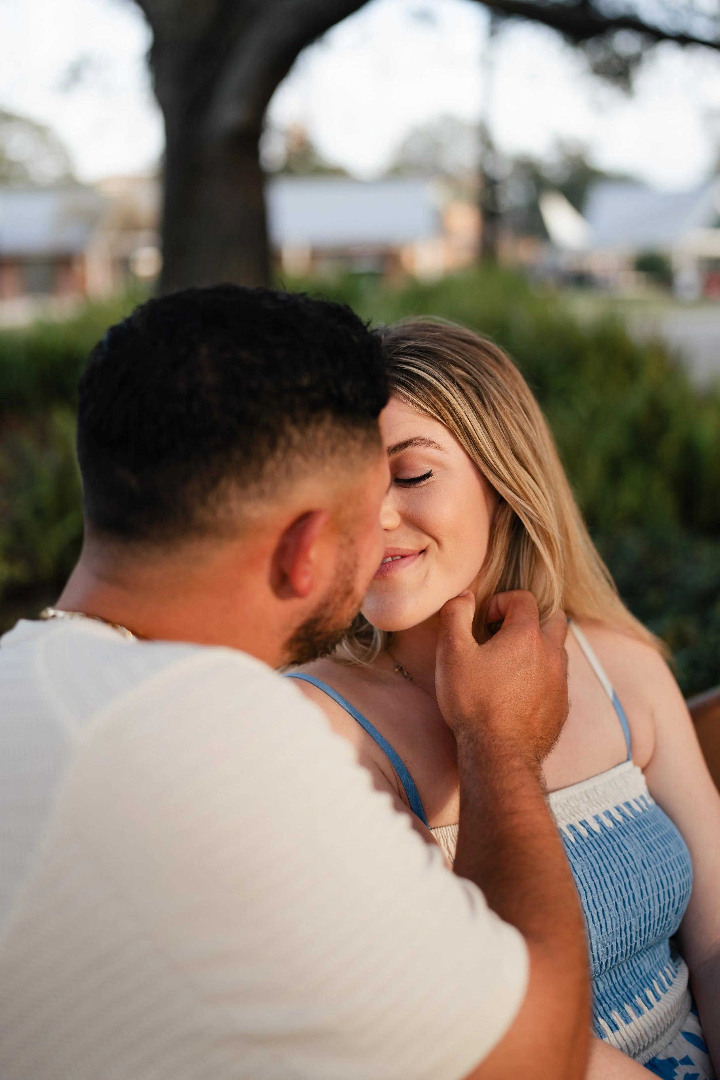 A young man gently touching a young woman's chin as they share an intimate moment outdoors, with trees and houses visible in the background.