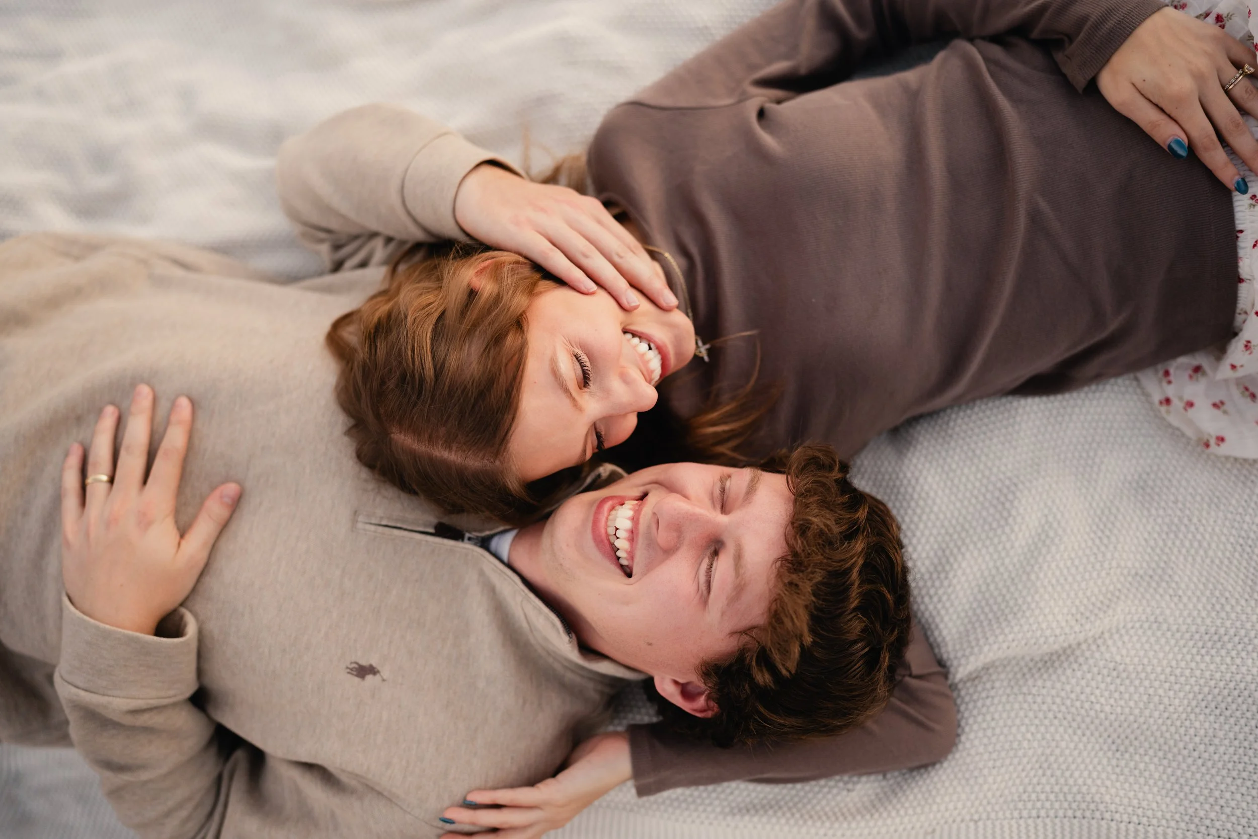 A young couple lying on a bed, smiling, with the woman resting her head on the man's chest and the man holding her close, sharing a joyful moment.