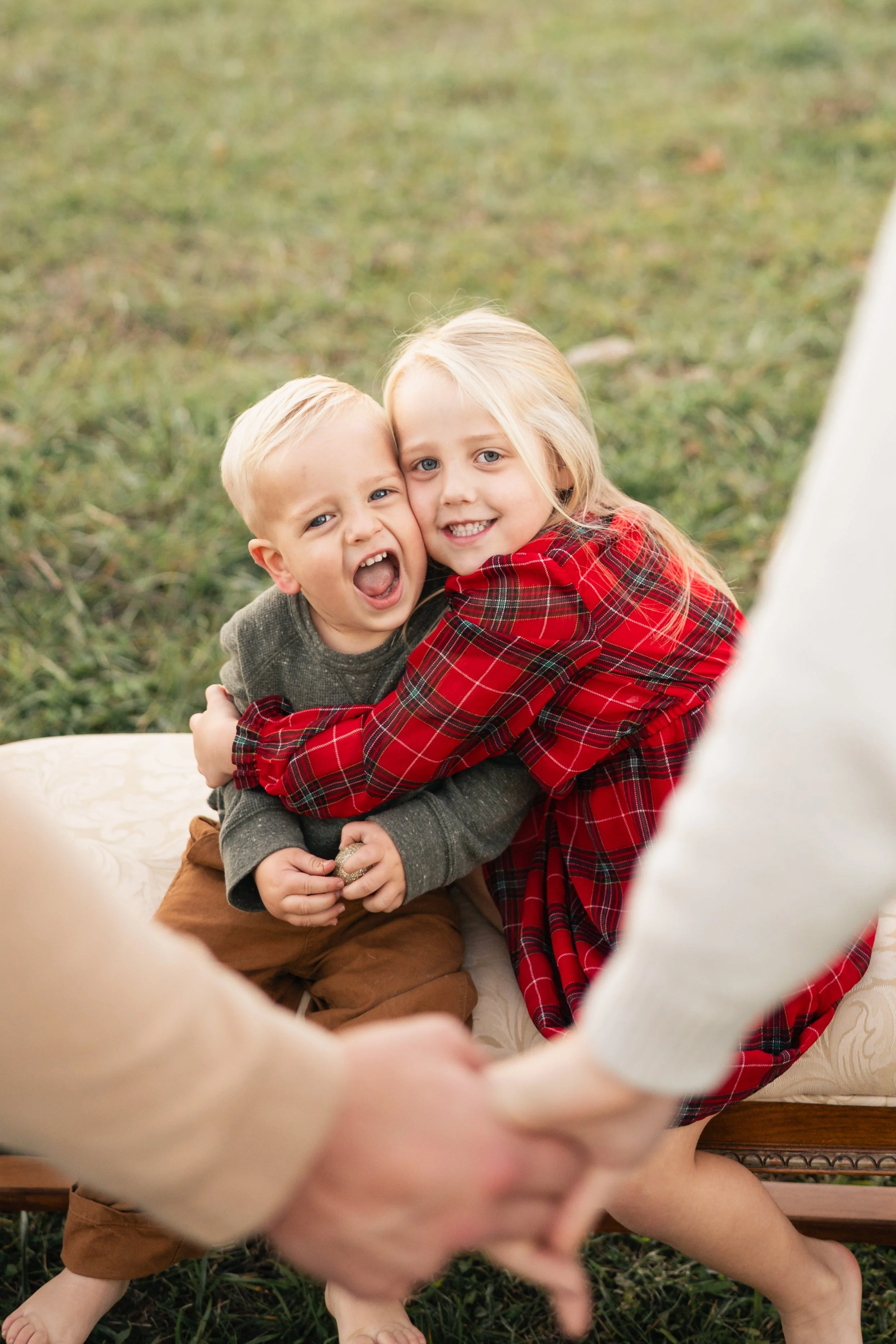 Two young children hugging and smiling, with a person in the foreground holding their hands, outdoors on a grassy area.