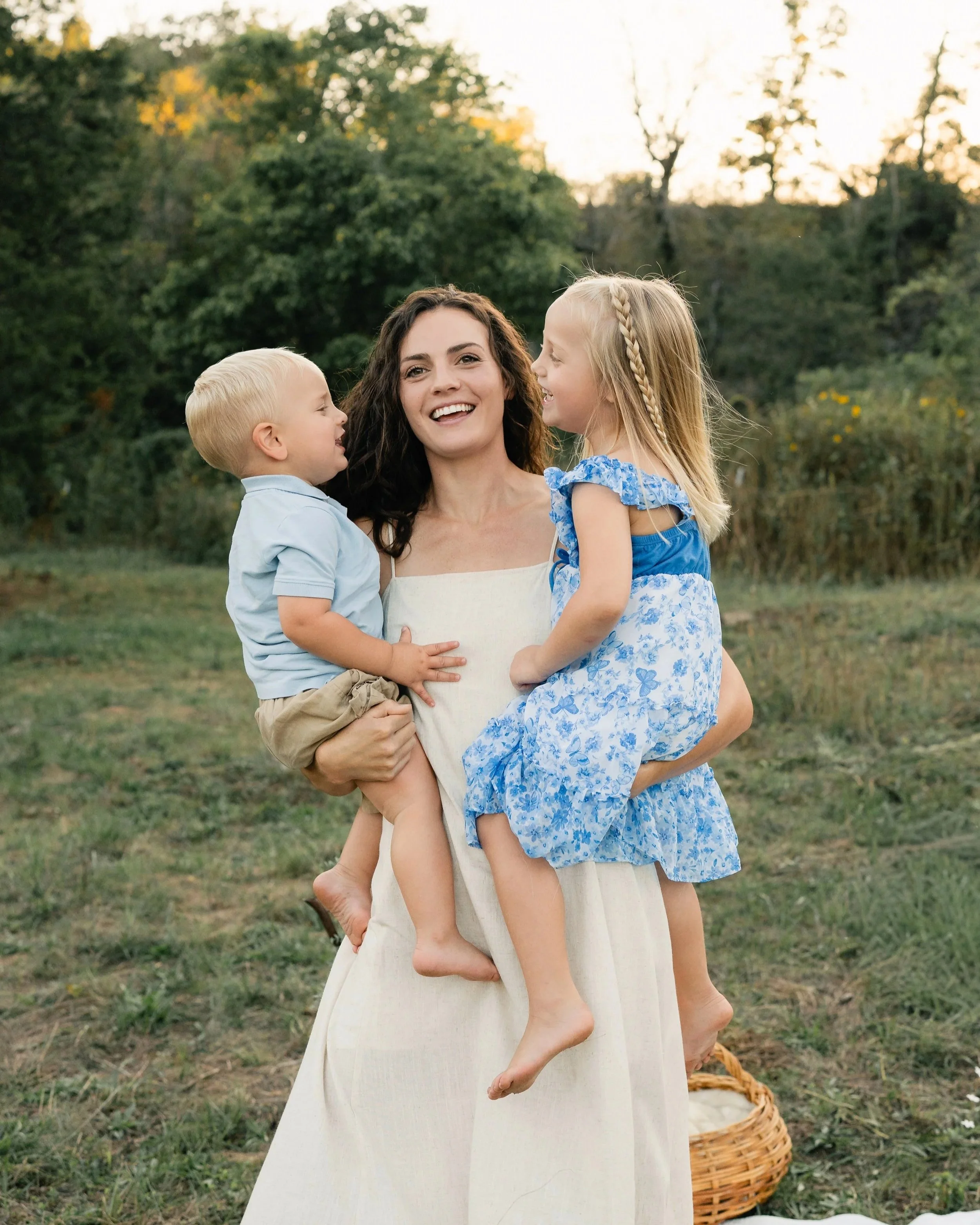 A woman smiling and holding two children outdoors in a grassy field with trees in the background during sunset.