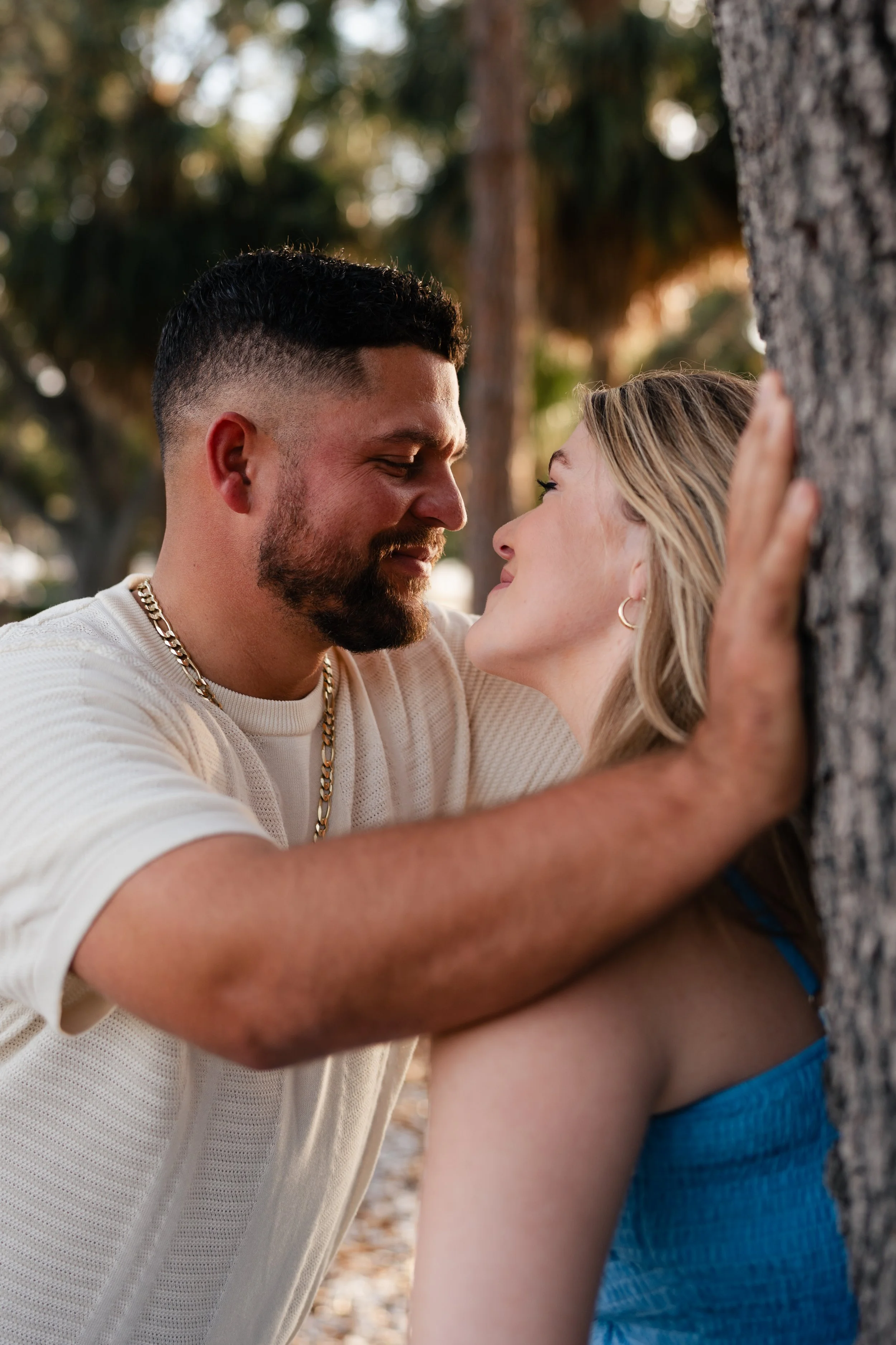 A man and woman face each other closely, smiling and leaning against a tree in a park, with sunlight filtering through the trees in the background.