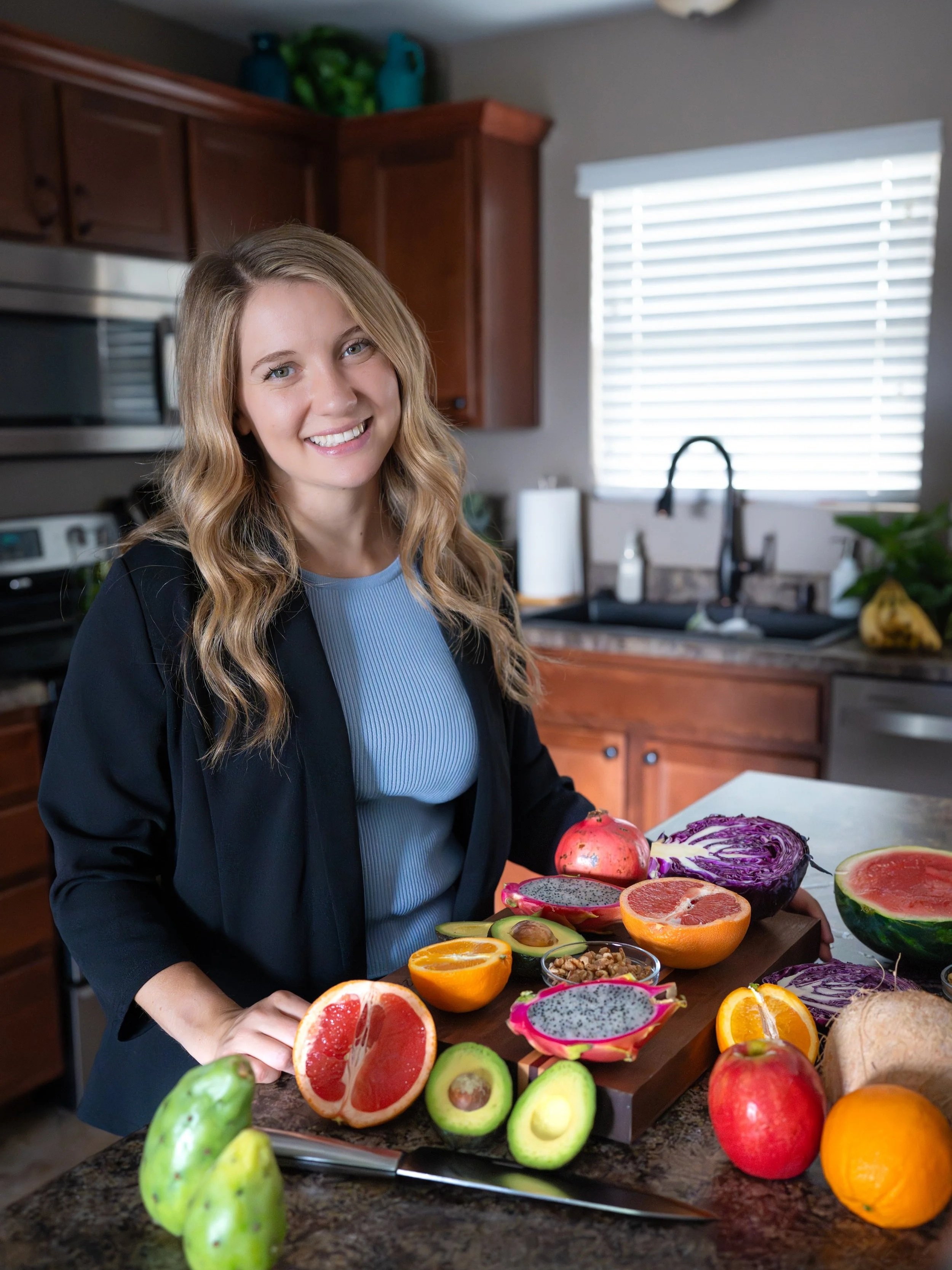 A woman with long wavy blonde hair and a blue shirt stands in a kitchen, smiling, surrounded by various sliced fruits on a chopping board and on the counter, including grapefruit, avocado, dragon fruit, and oranges.