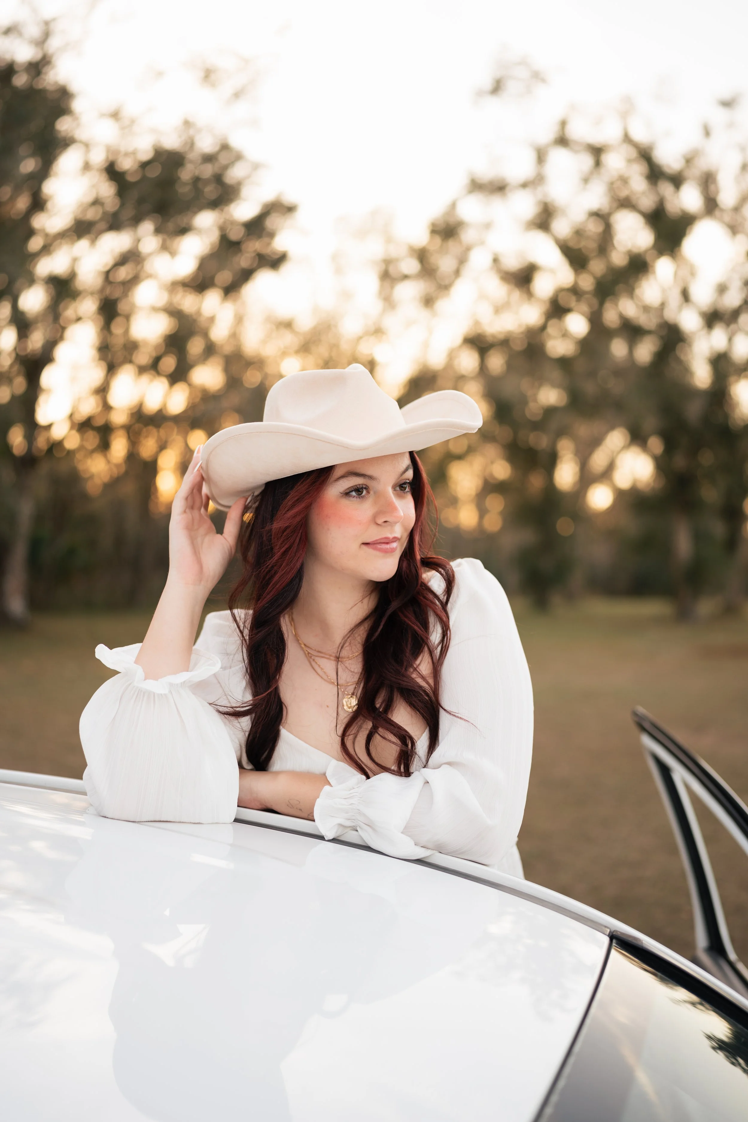 A young woman with auburn hair wearing a white cowboy hat and white blouse, leaning on the roof of a car outdoors during sunset.