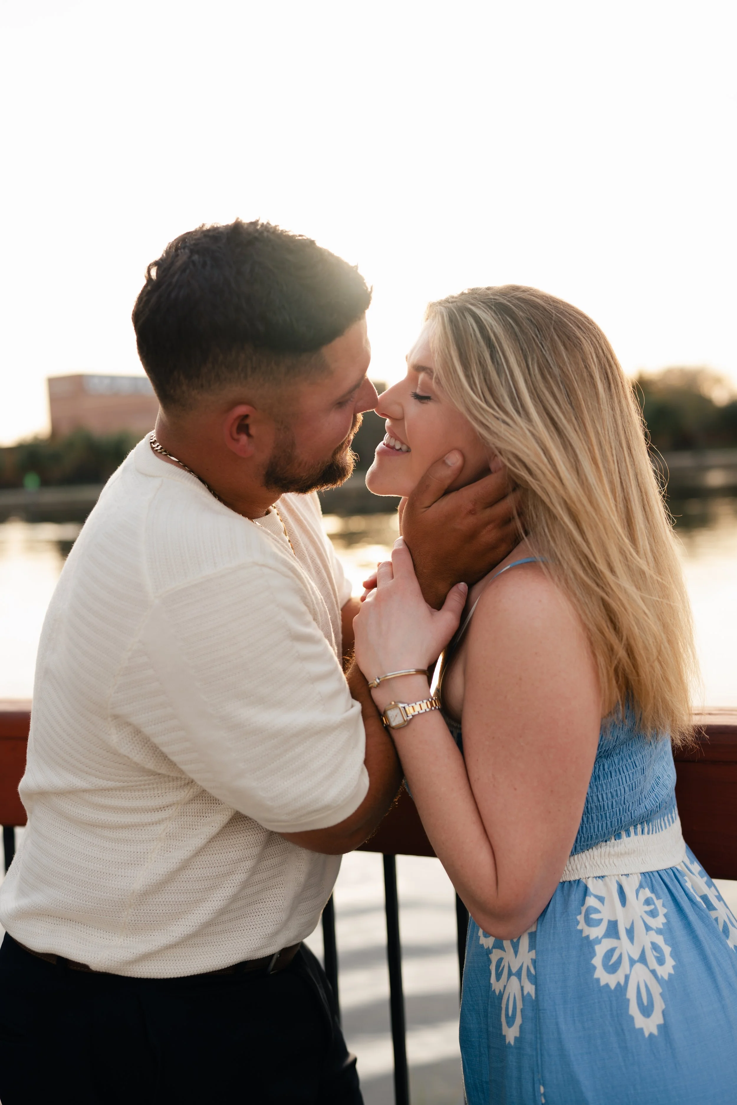 A couple embraces tenderly by a river, with a sunset in the background.