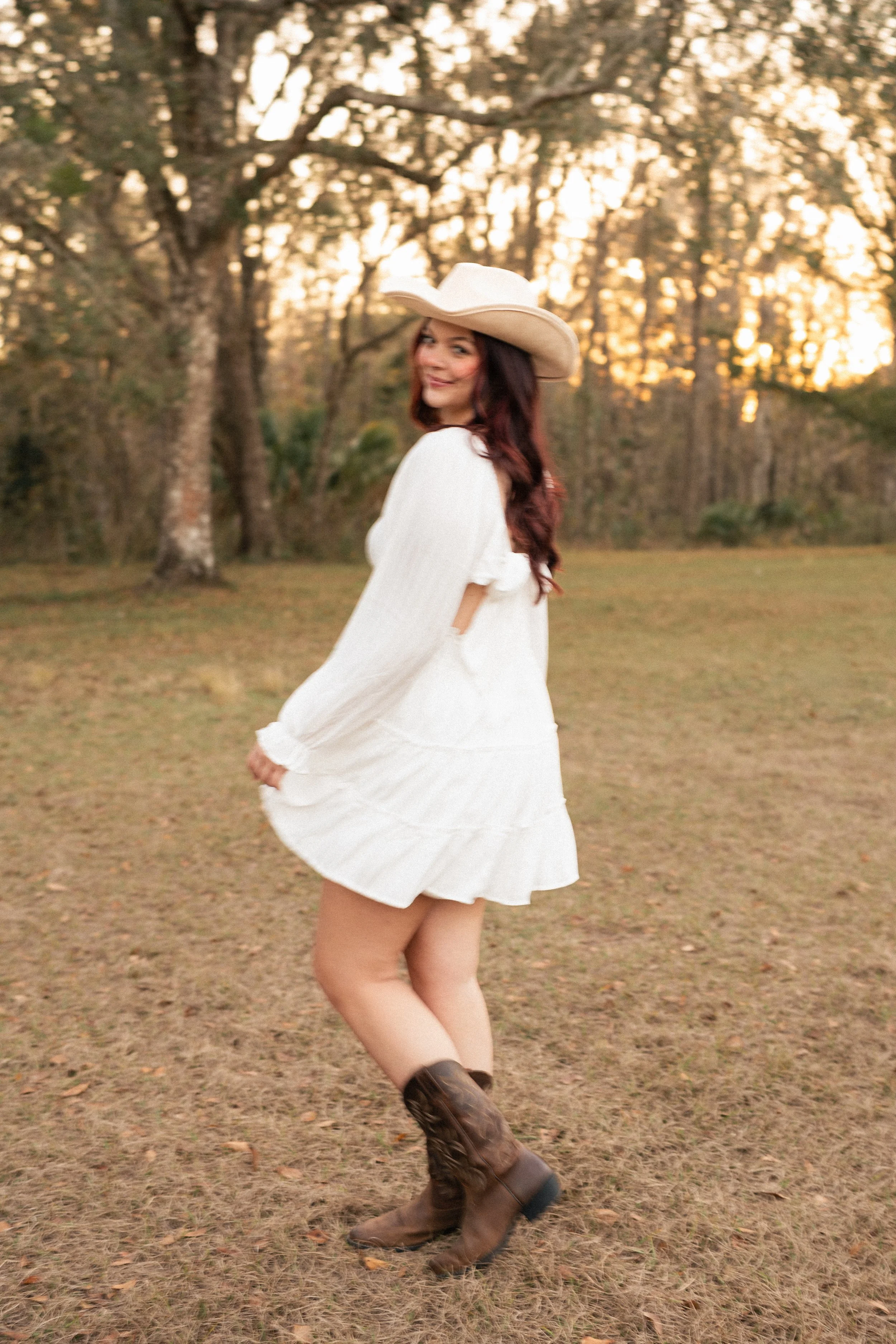 A woman in a white dress and cowboy boots, wearing a cowboy hat, standing outdoors near trees during sunset.