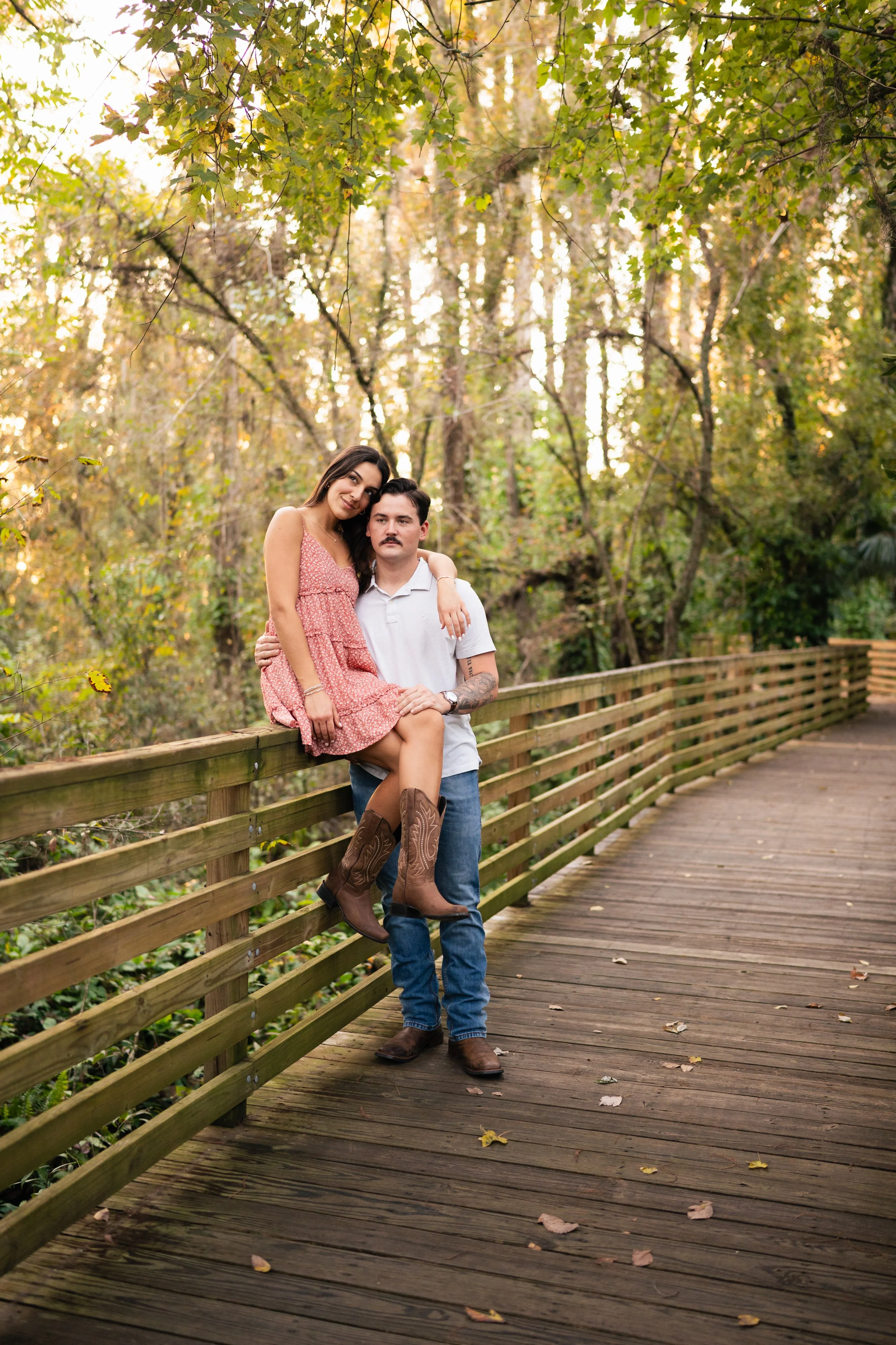 A couple on a wooden bridge in a forest during autumn, with the woman sitting on the railing and the man standing beside her.
