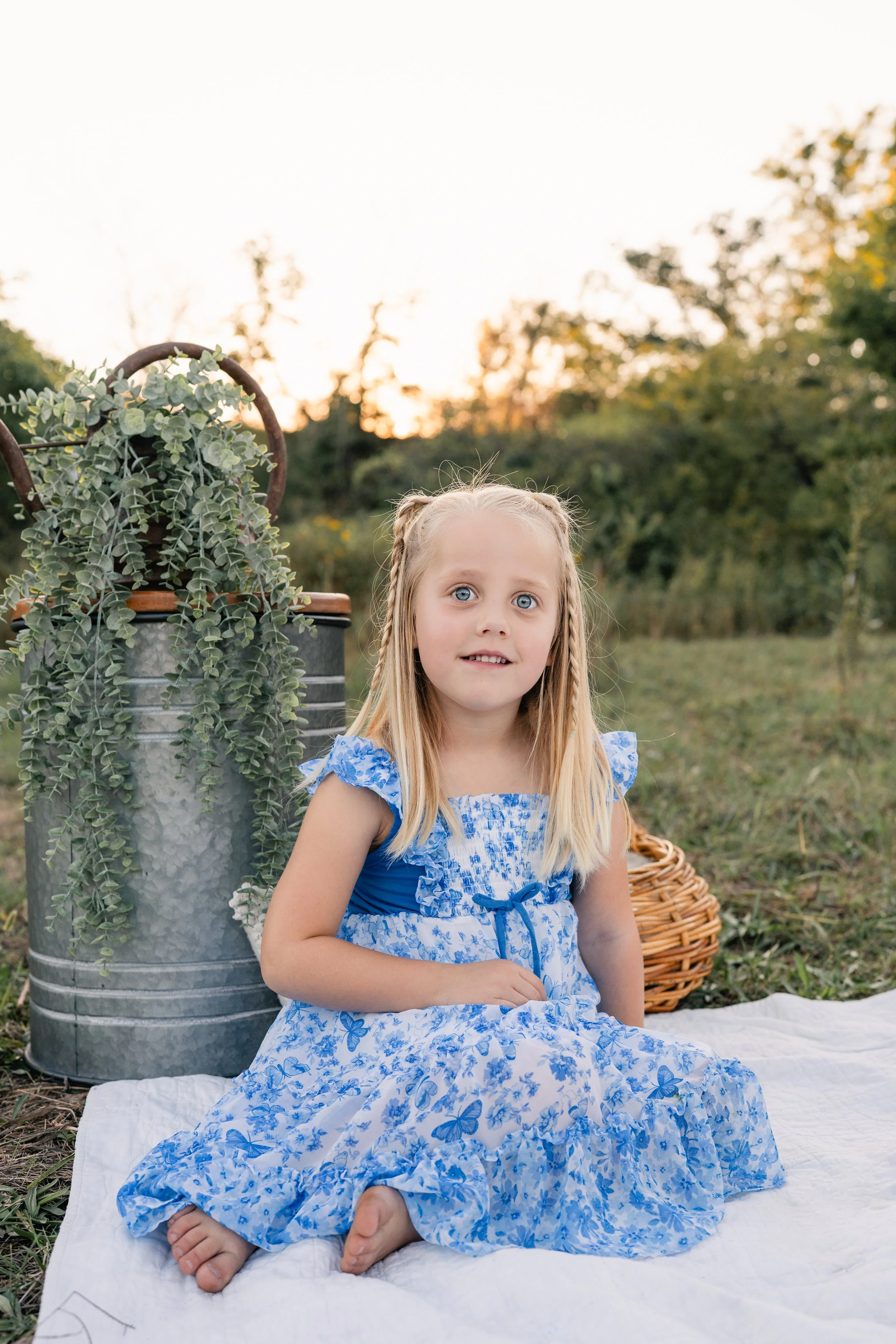 A young girl with blonde hair and blue eyes sitting on a white blanket outdoors during sunset, wearing a blue and white floral dress, next to a metal watering can with a green plant, and a wicker basket in the background.
