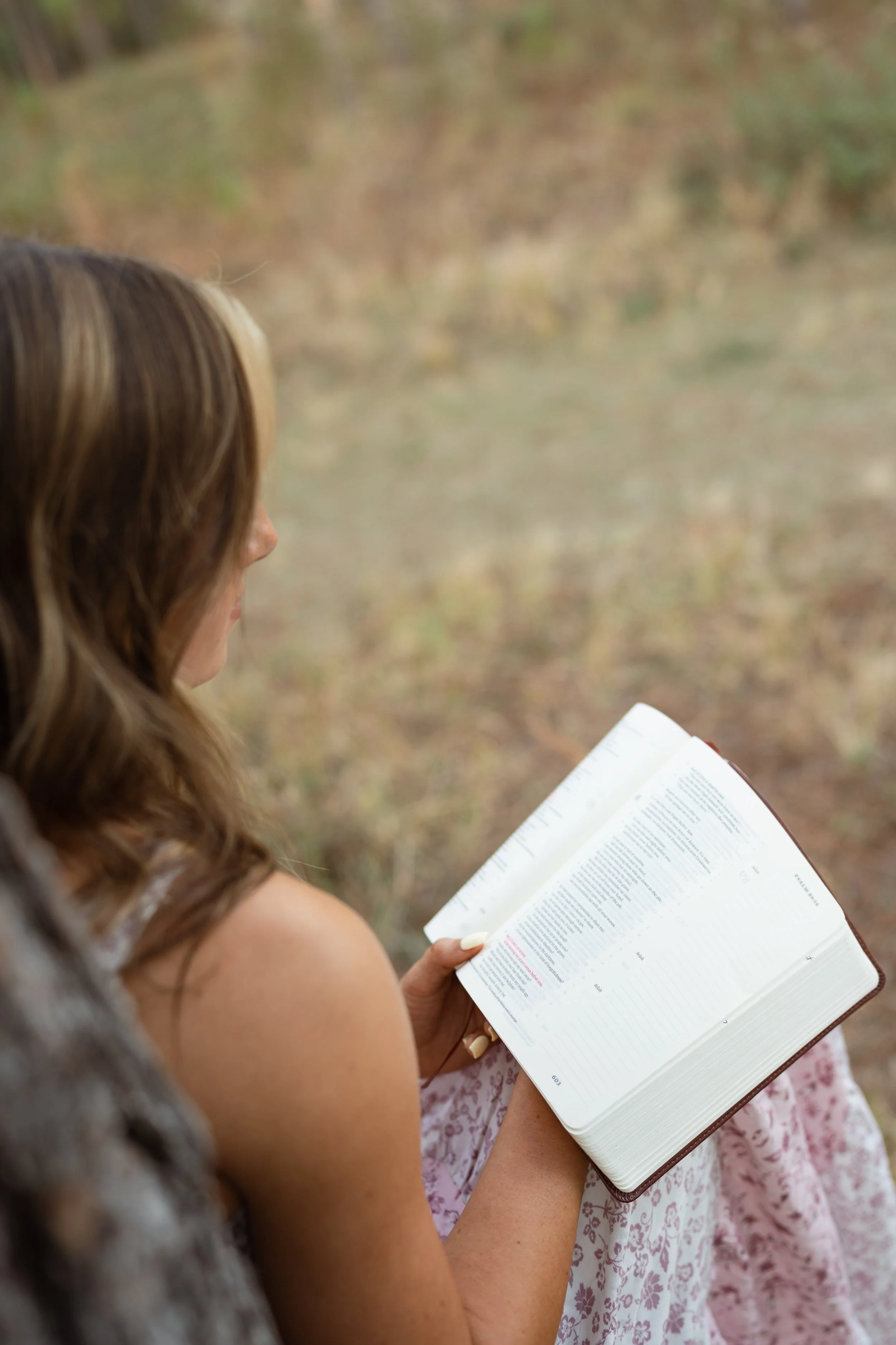 A woman with wavy brown hair and a sleeveless top sitting outdoors, reading a white-lined book with text, with a blurred natural background.