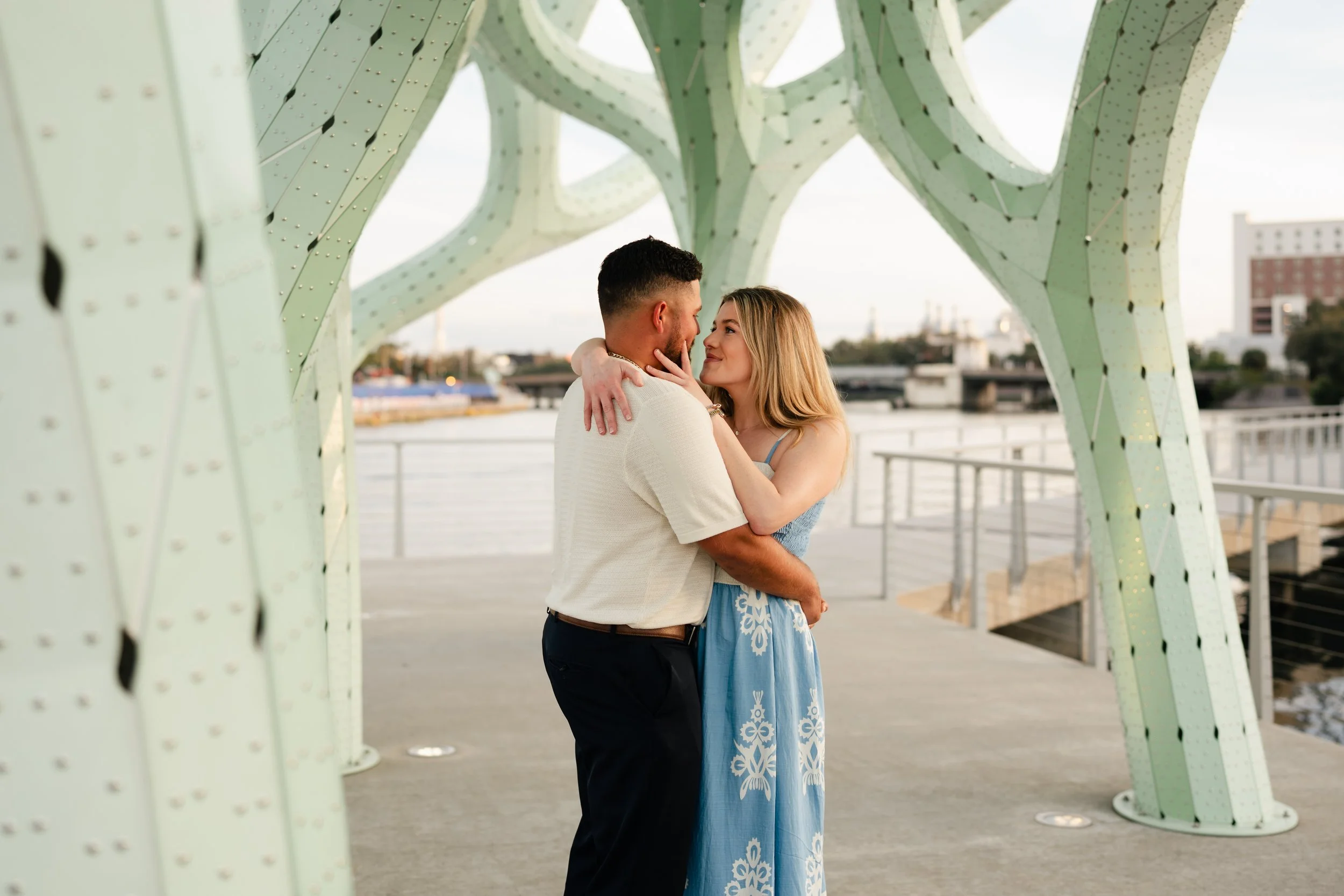 A couple embraces under a green modern architectural structure by the water during sunset.