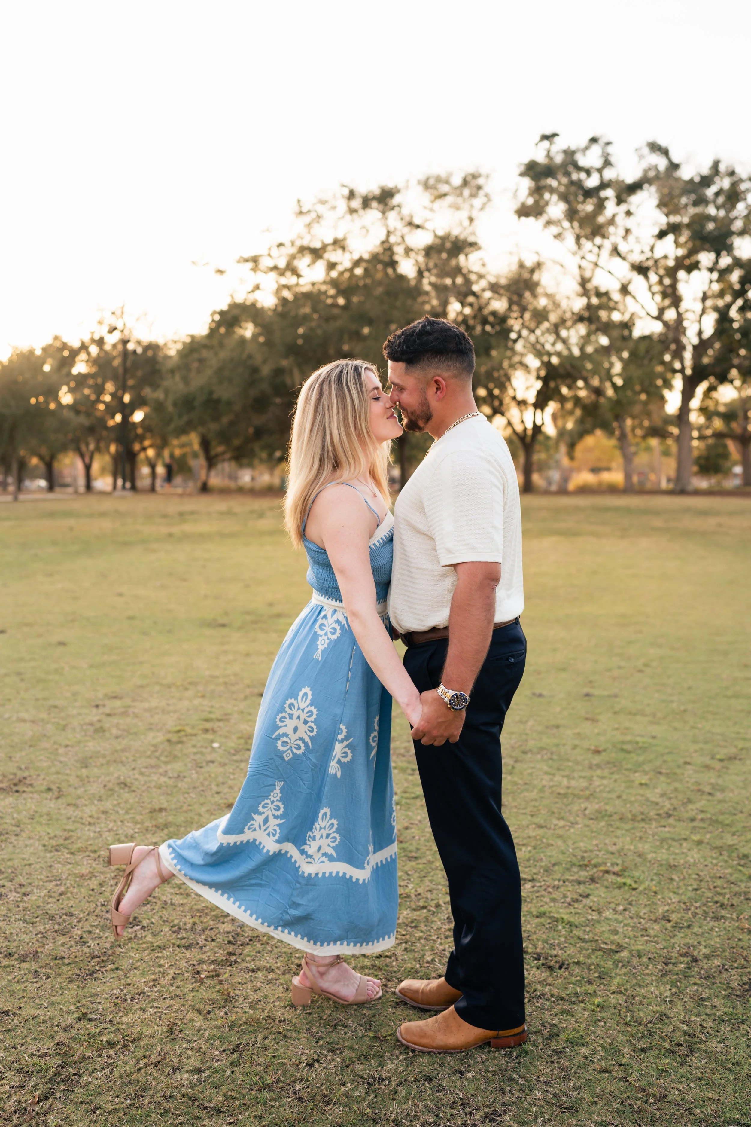 A couple standing close together, holding hands, about to kiss outdoors in a park during sunset, with trees in the background.