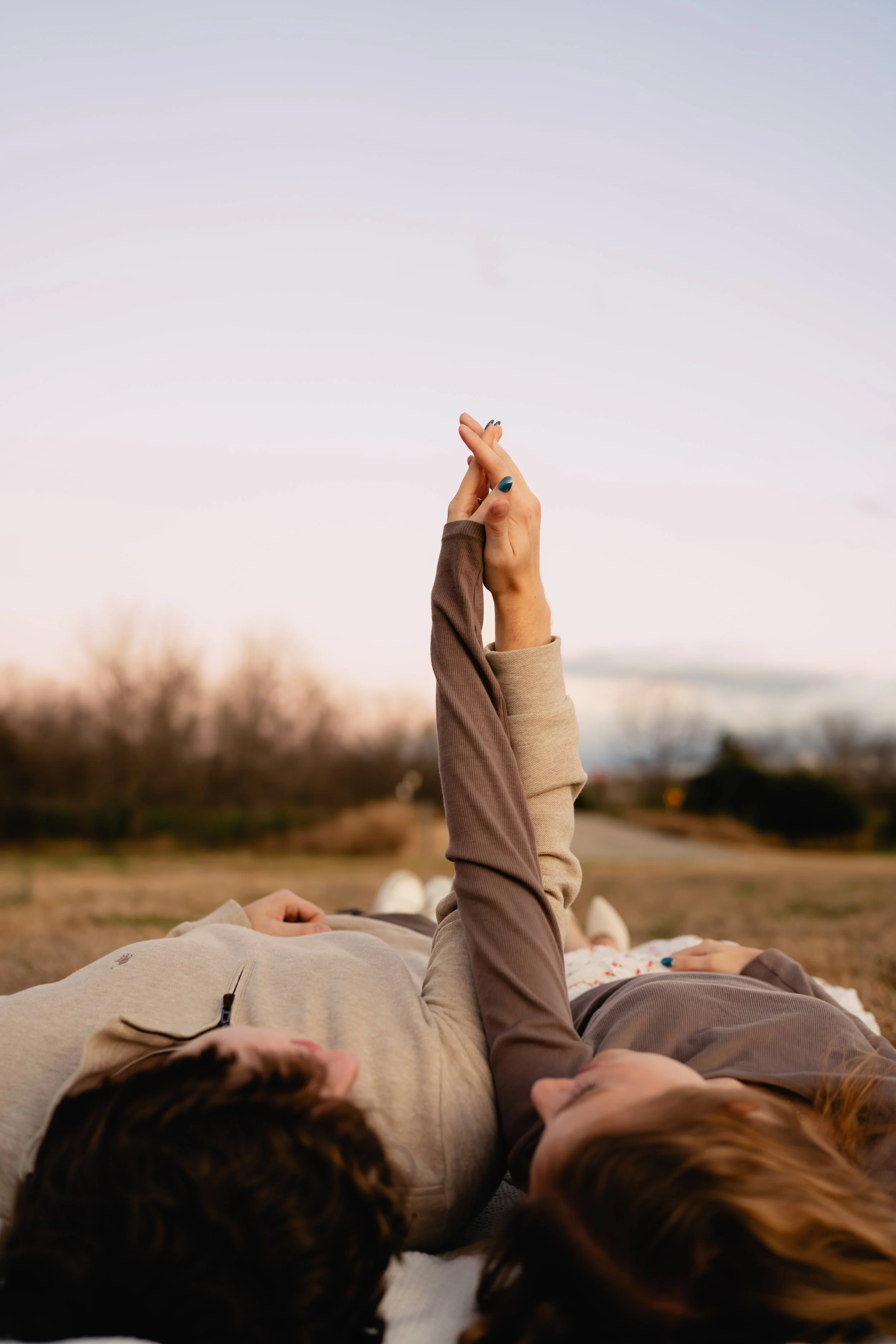 Two women are lying on their backs outdoors, holding hands, with one woman holding an upward hand gesture against a sunset sky.