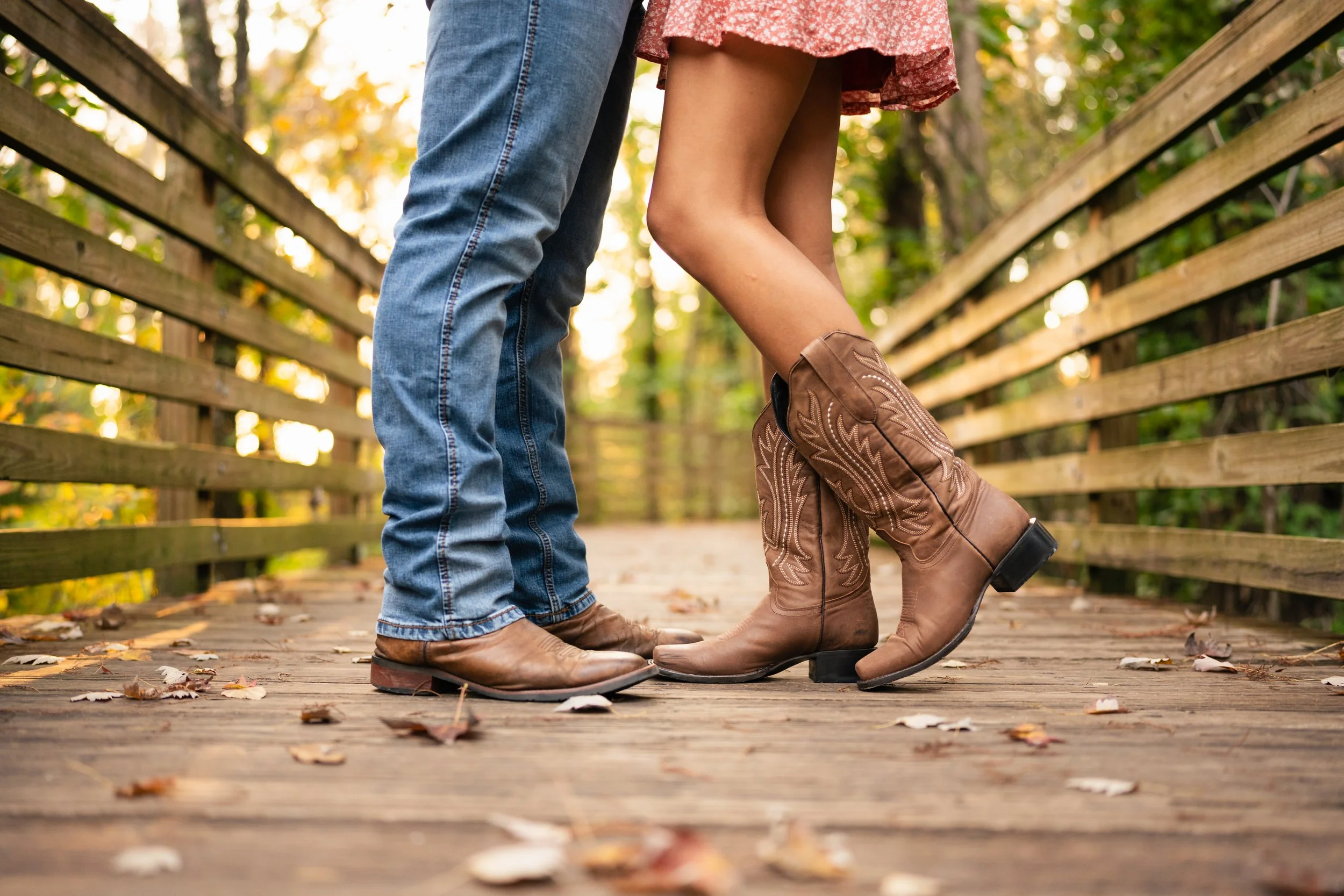 Close-up of a couple's legs and feet standing on a wooden bridge, with the woman wearing cowgirl boots and the man wearing cowboy boots and jeans, surrounded by autumn foliage.