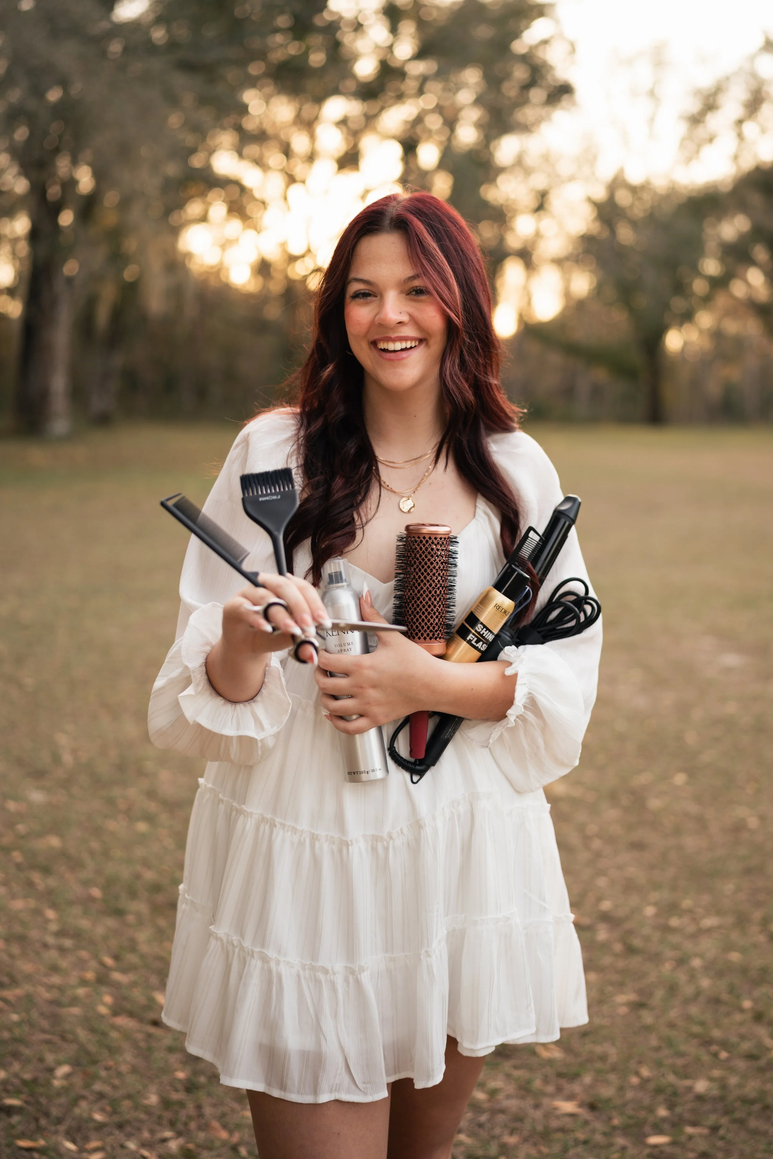A smiling woman in a white dress holding various hair styling tools and products outdoors during sunset.