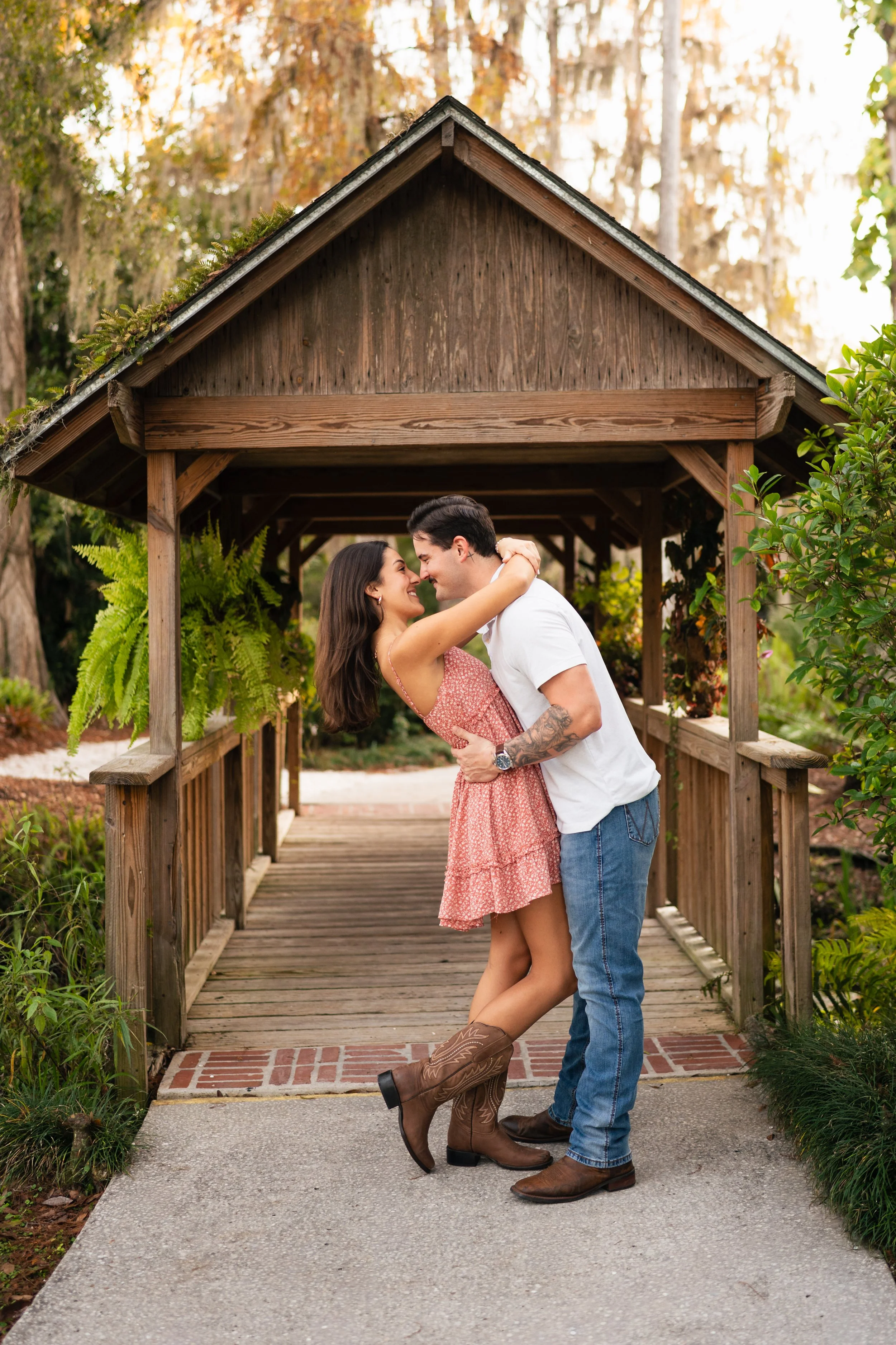 A couple embracing and smiling in front of a wooden pavilion, with greenery and trees around.