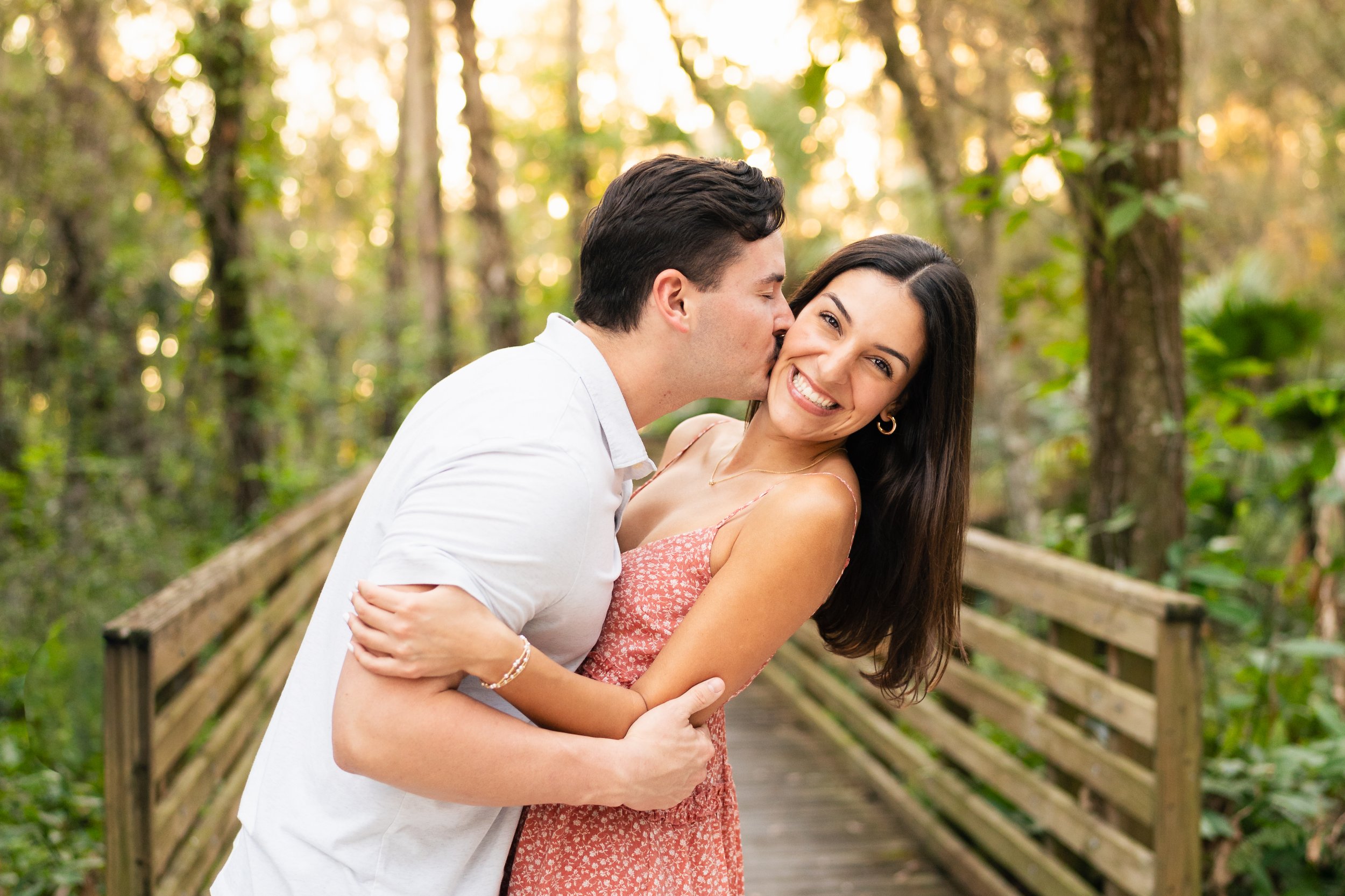A couple sharing a kiss and smiling on a wooden bridge in a forest during daytime.