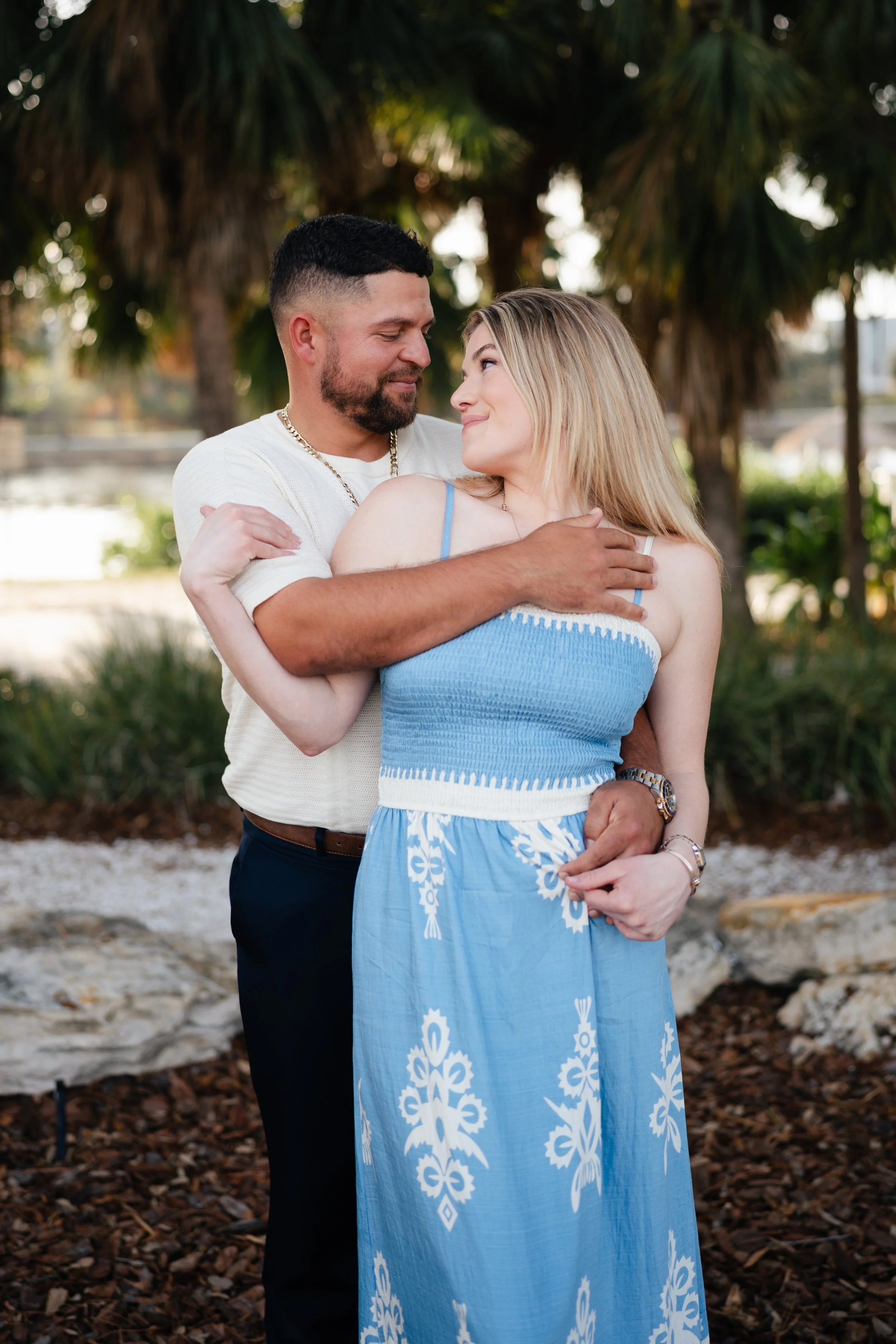 A couple embracing outdoors in front of palm trees, dressed in casual summer clothing, smiling at each other.